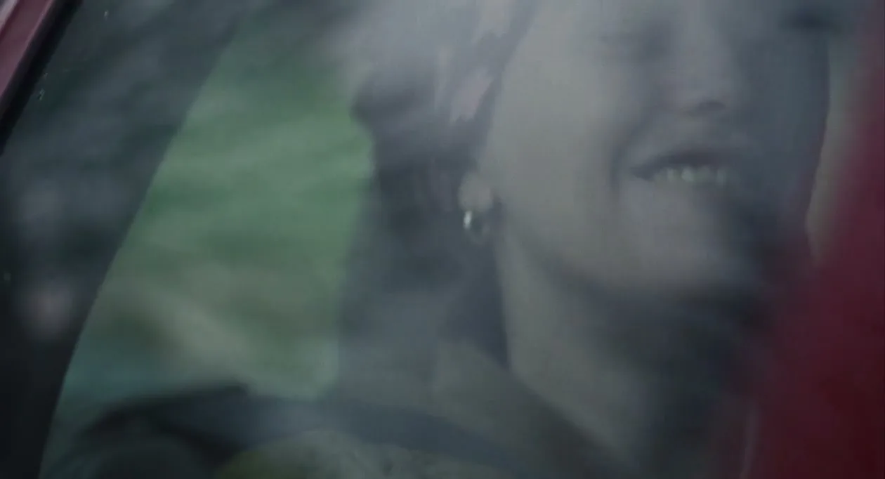 A woman with dark hair and a patterned headscarf smiles, seen through a car window, with a green background.