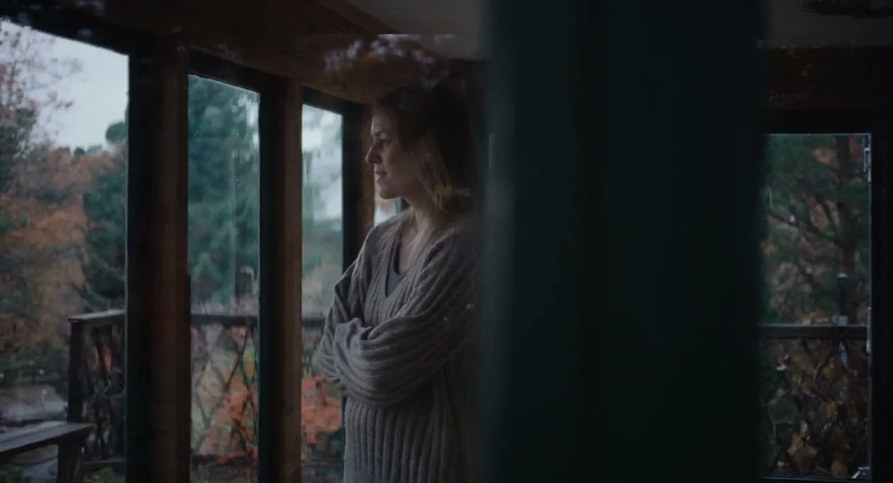A woman with blonde hair, wearing a beige sweater, stands with her arms crossed, looking out a window at a rainy autumn landscape.