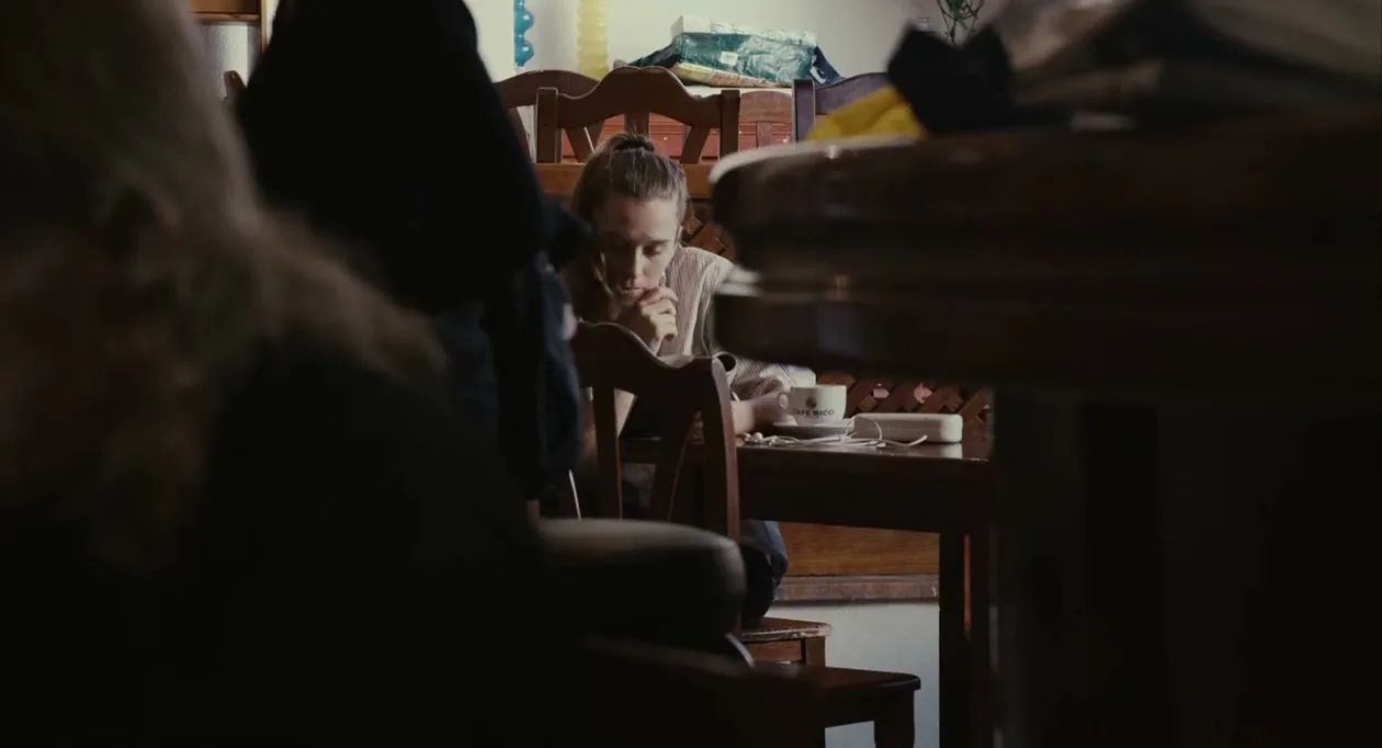 A young woman with long hair sits at a wooden table, looking down thoughtfully, with a cup and phone nearby in a dimly lit room.