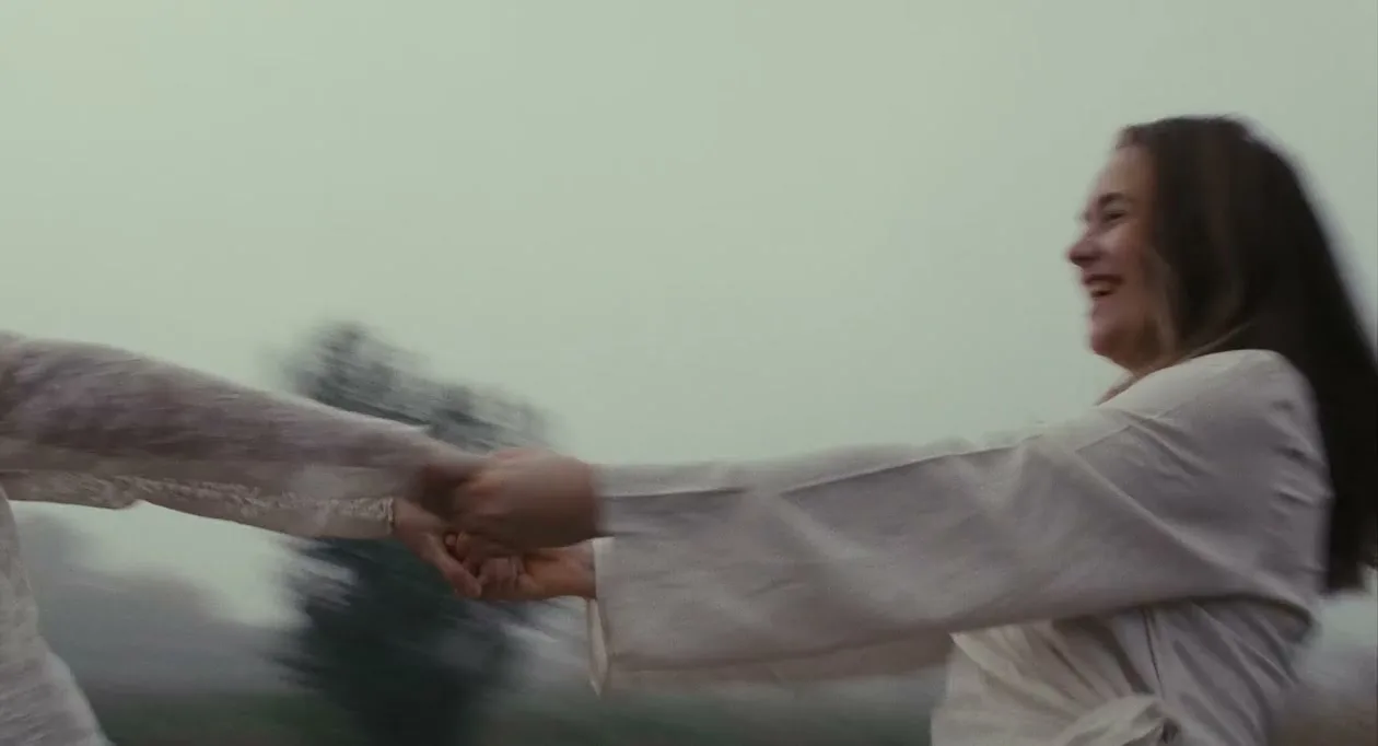 Two women in white dresses hold hands and spin in a circle, laughing joyfully in a foggy outdoor setting.
