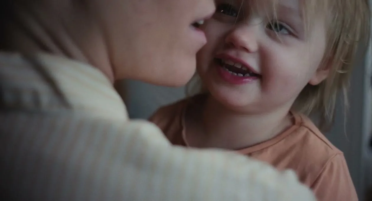 A mother whispers to her smiling toddler, who has blonde hair and is wearing an orange shirt, in a close-up shot.