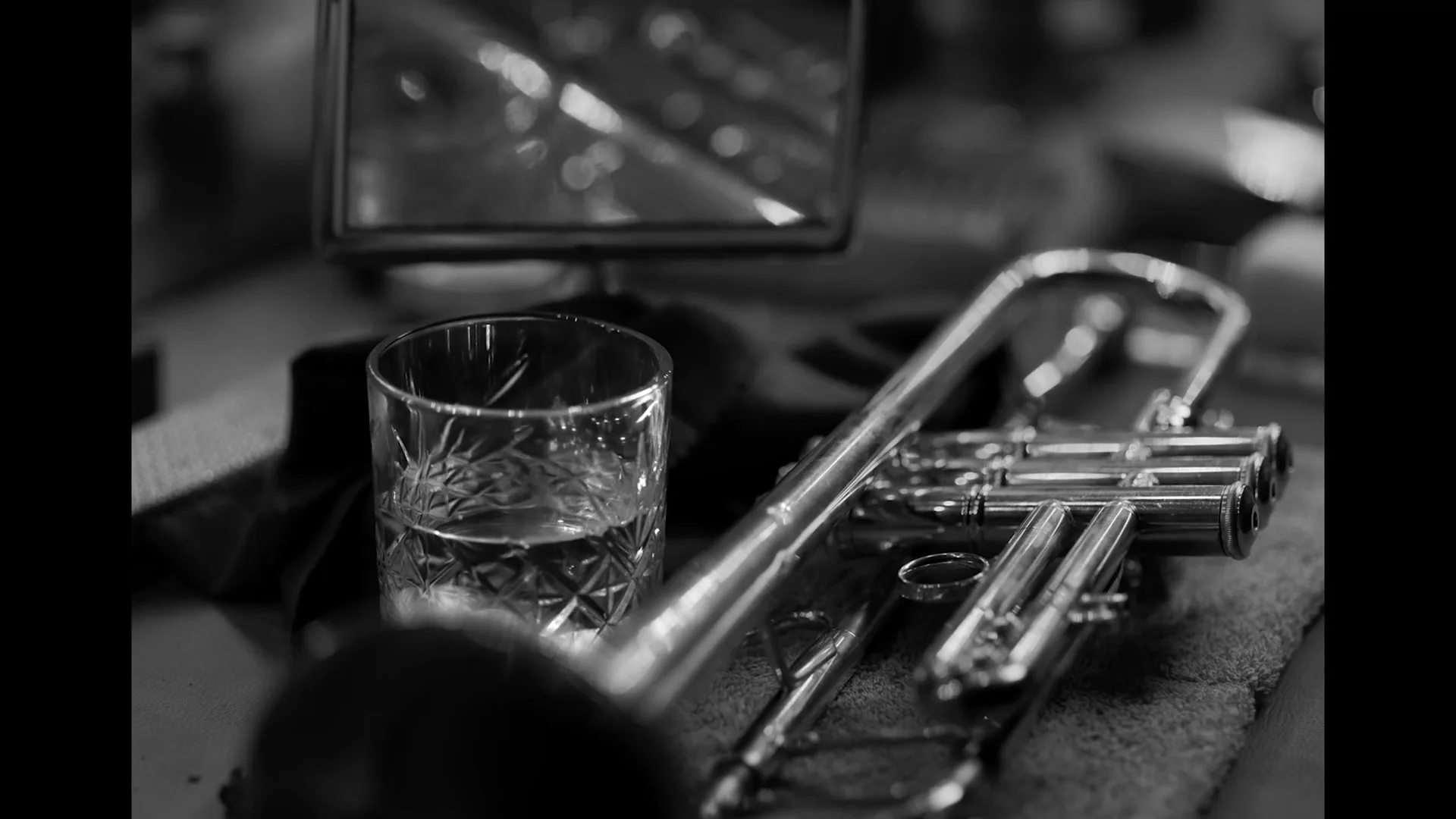 A close-up, black and white shot of a trumpet and a glass of liquid resting on a surface, with a small mirror in the background.