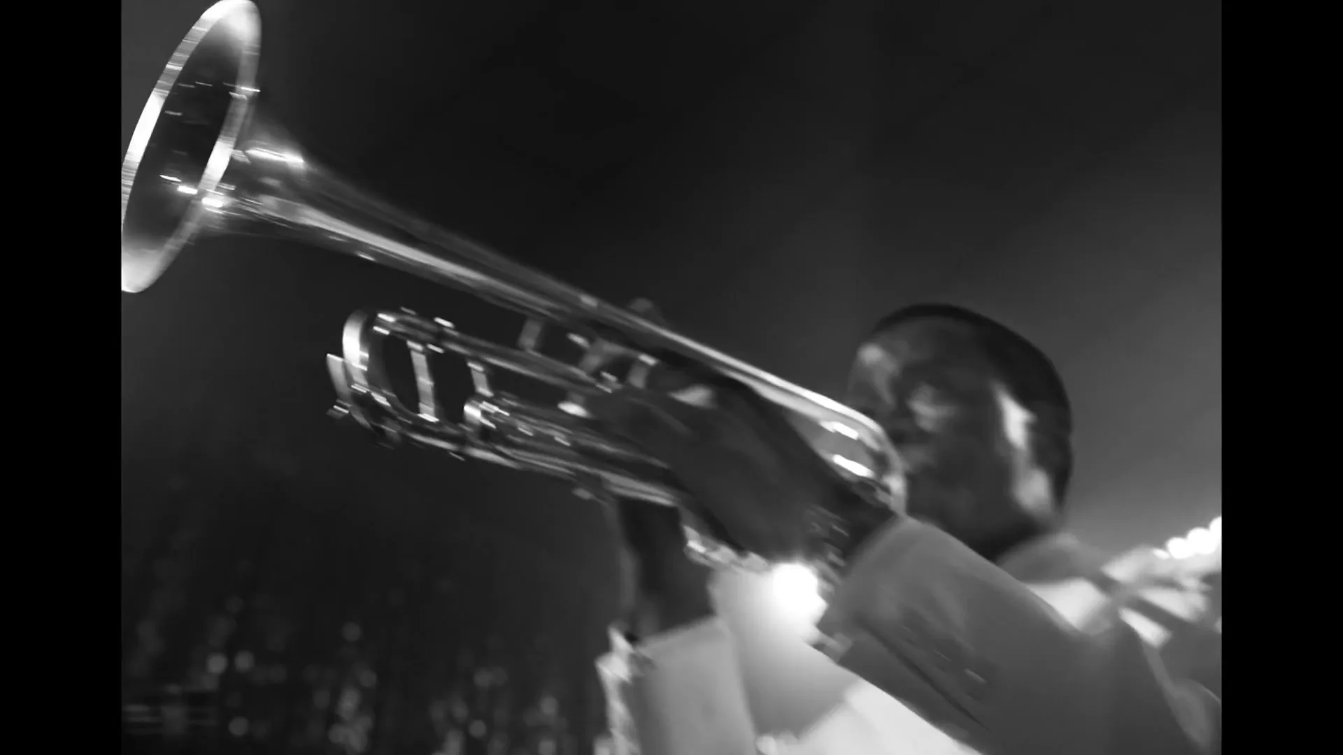 A black and white shot of a male musician in a white suit playing a trumpet on a stage, with the instrument reflecting light.
