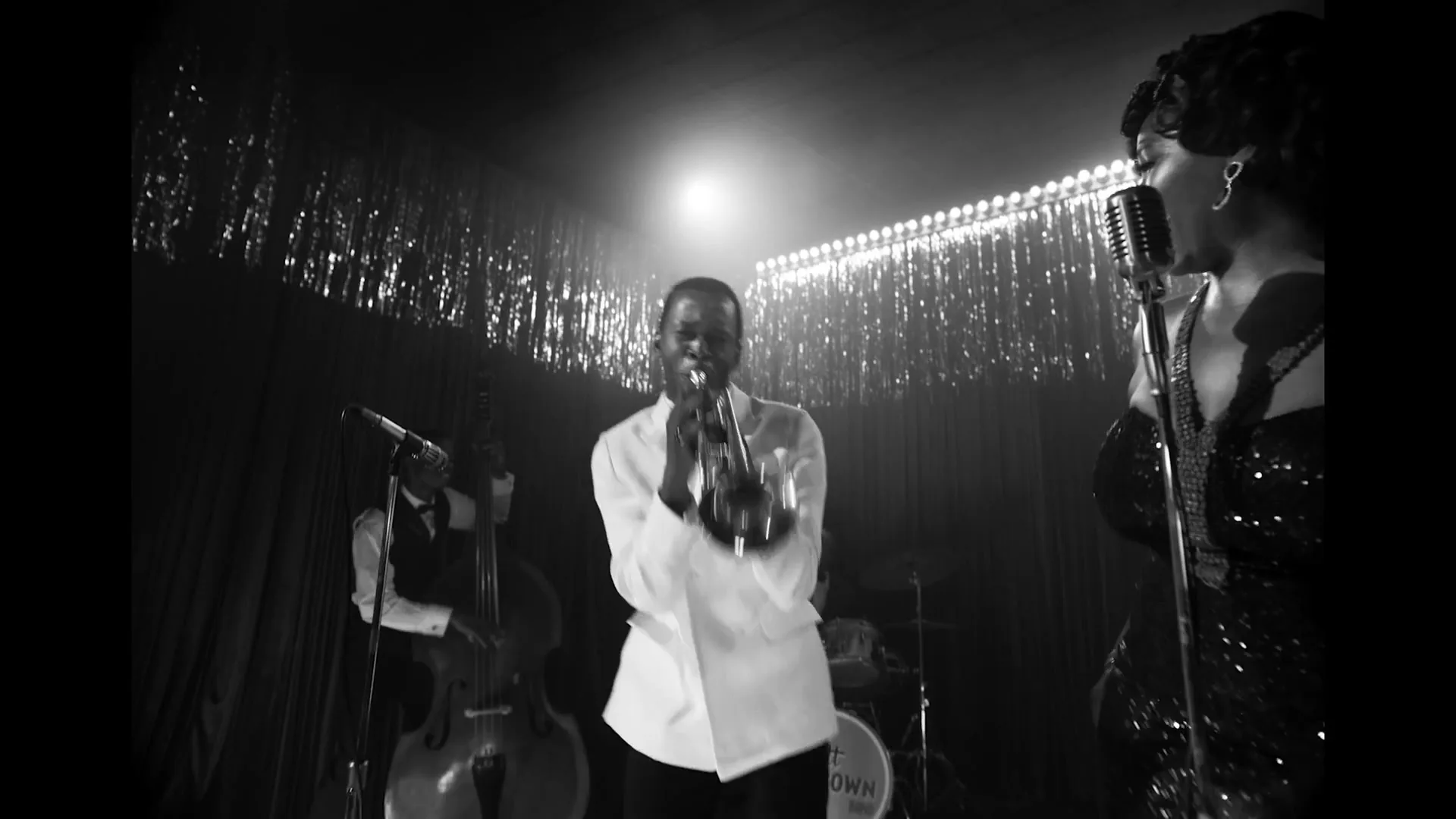 A Black male trumpet player in a white suit performs on stage with a female singer and a bassist in a dimly lit club.