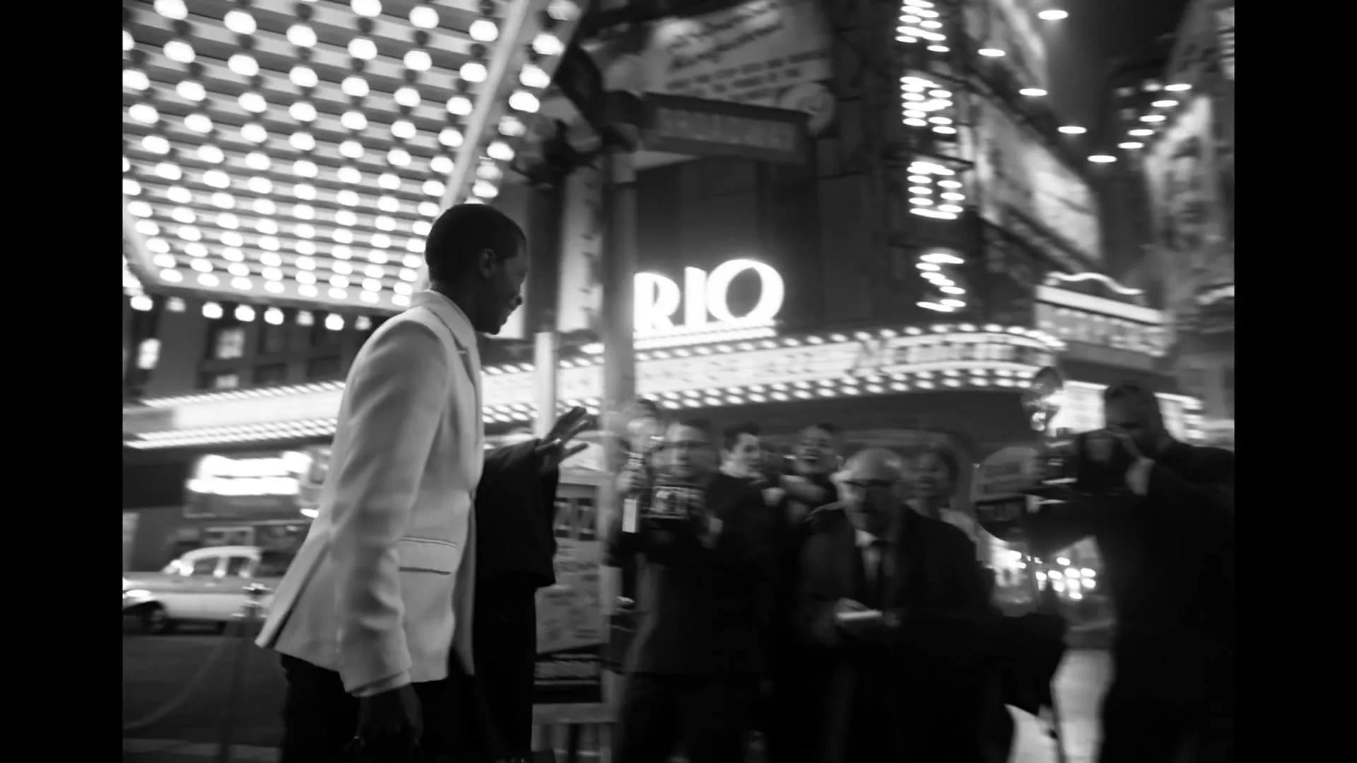 A man in a white suit waves to a crowd of photographers and fans as he exits a building onto a busy city street at night.