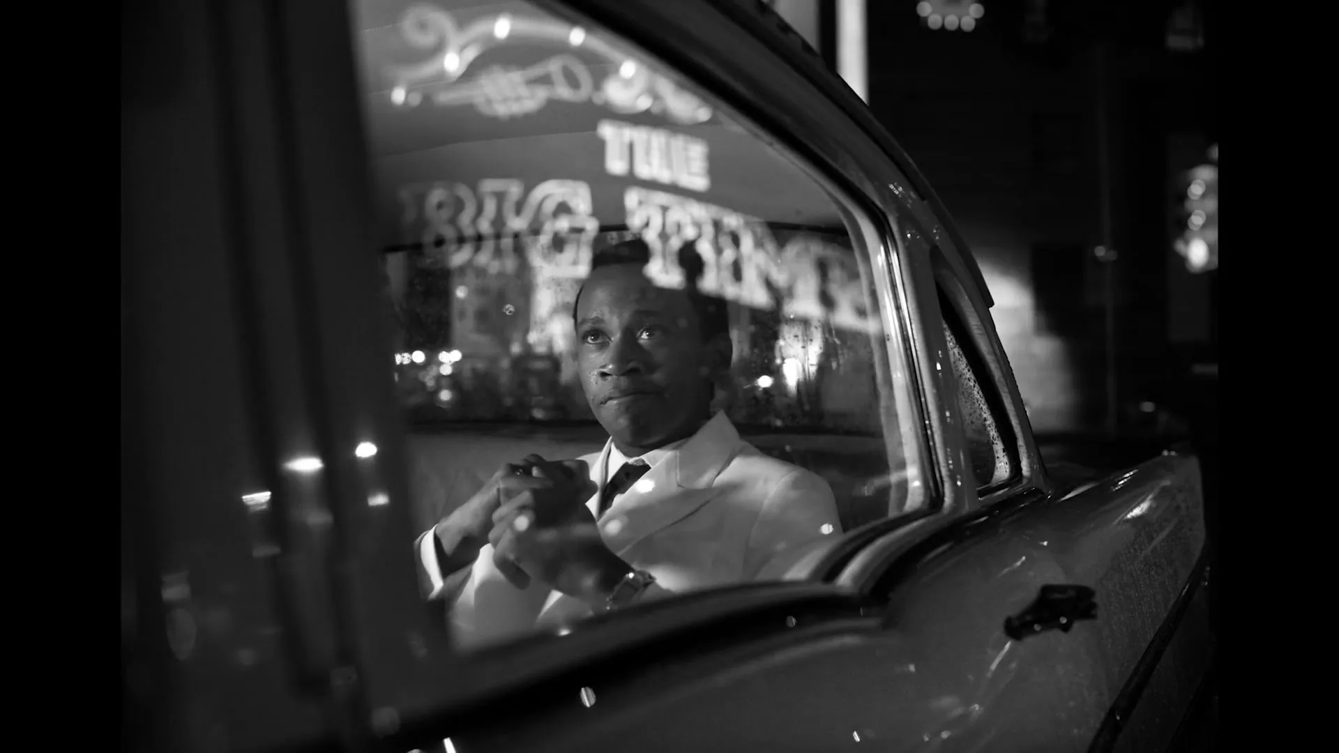 A young Black man in a white suit sits in the passenger seat of a car at night, looking out the window with city lights reflecting.