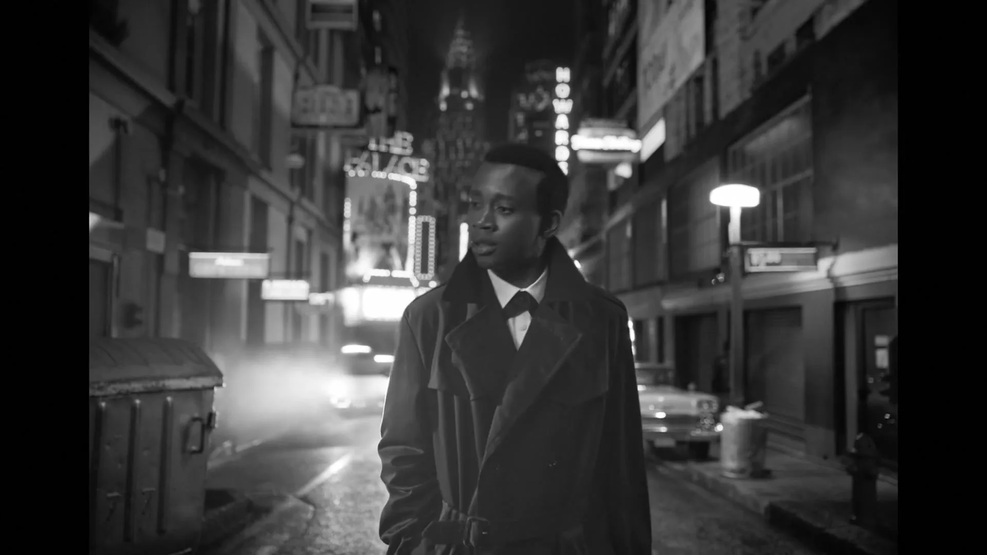 A young man in a trench coat walks down a dark, foggy city street at night, looking up at the neon signs.