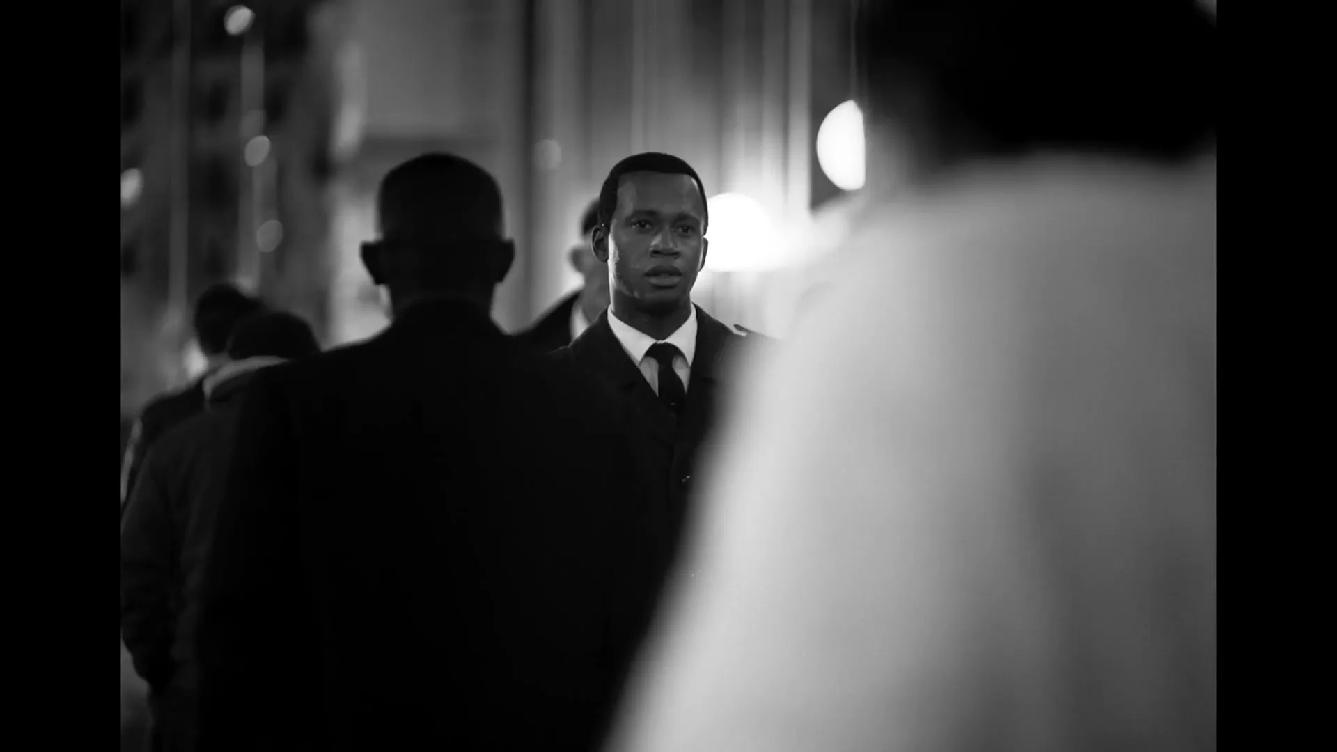 A young Black man in a trench coat and tie looks around nervously as he walks through a busy city street at night.