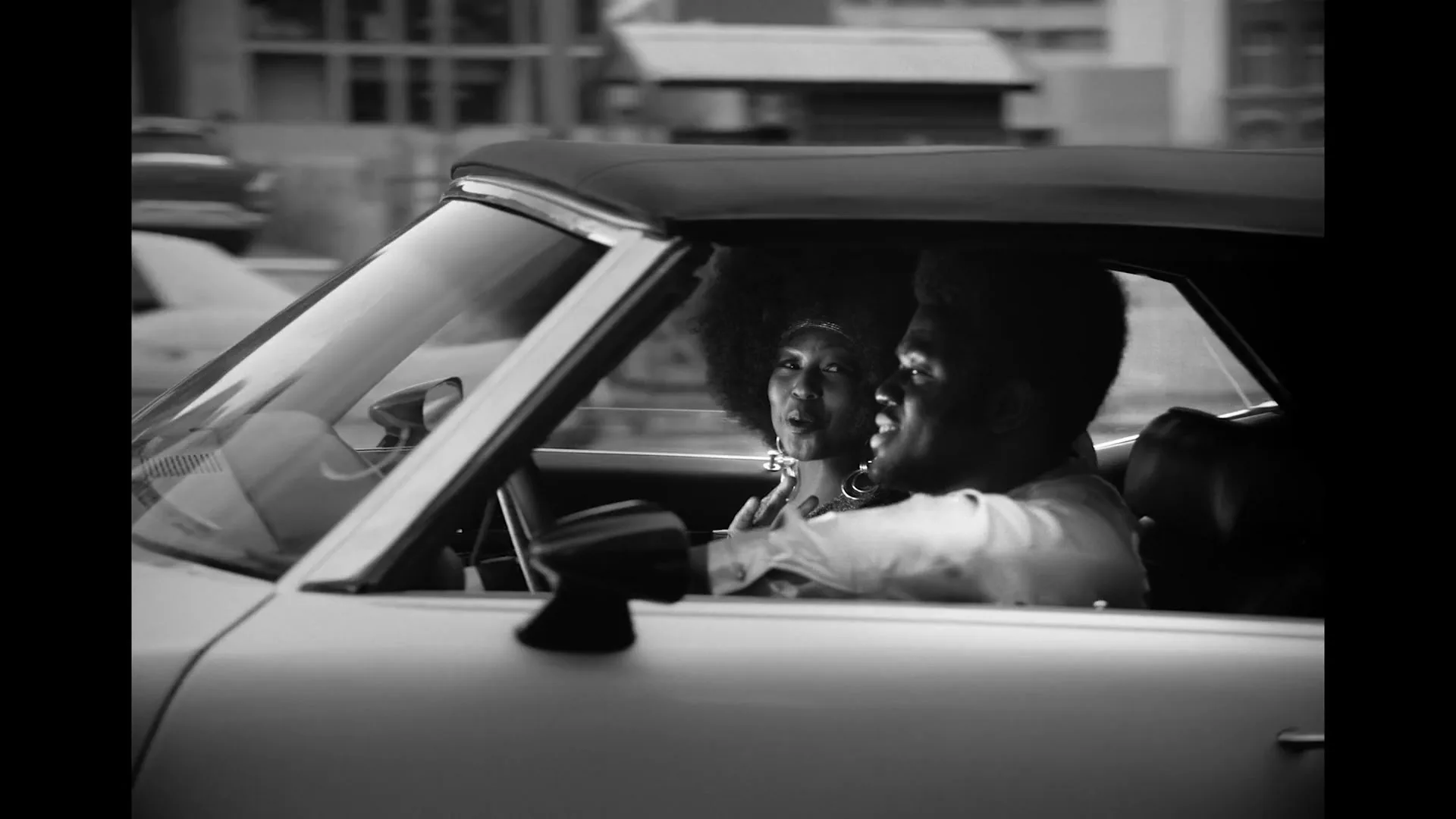 A Black couple in their 20s, dressed in casual attire, drives a convertible car down a city street, looking happy and engaged in conversation.