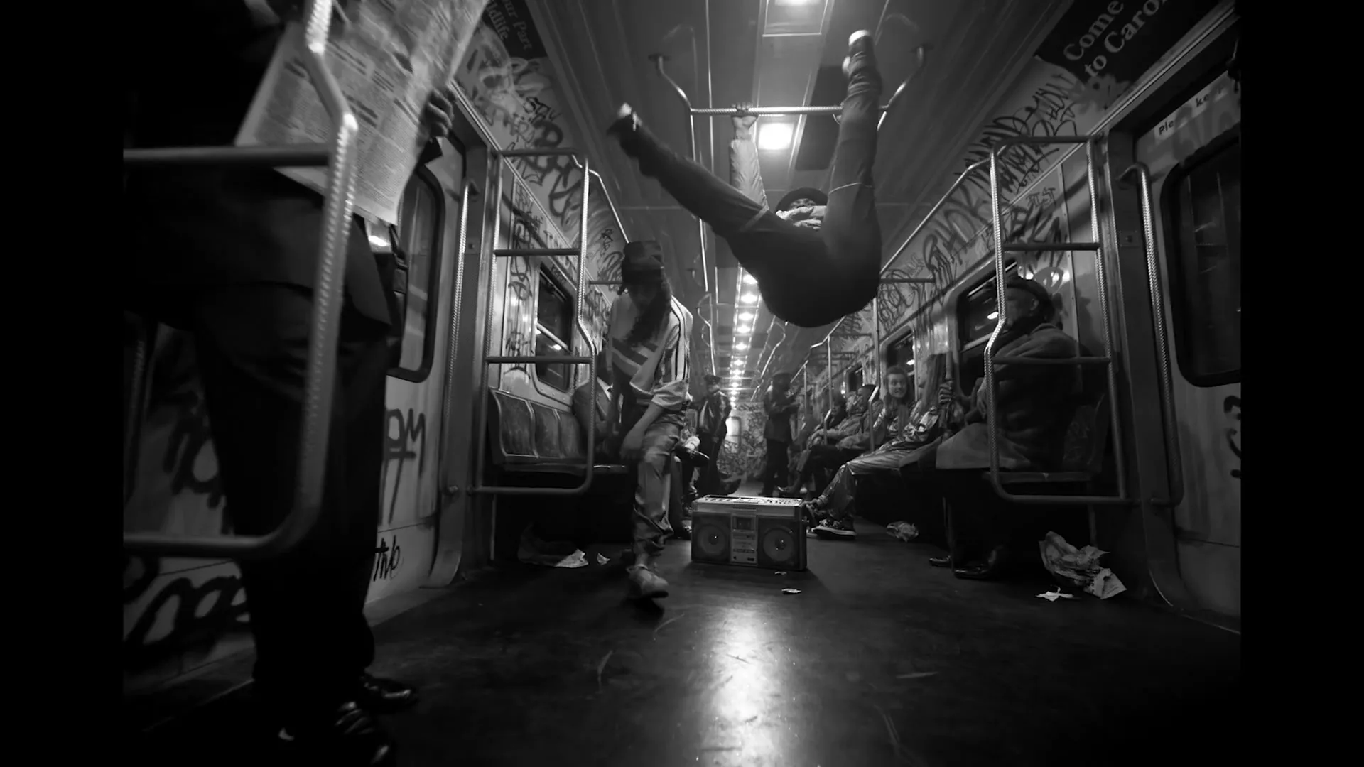A young man in a jacket hangs upside down from a subway car handrail, while other passengers sit or stand around him, in a black and white shot.