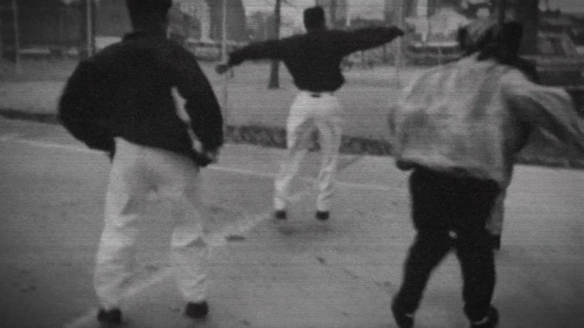 Three young men in casual clothes dance energetically on a paved surface, with a blurred urban background and graffiti wall.