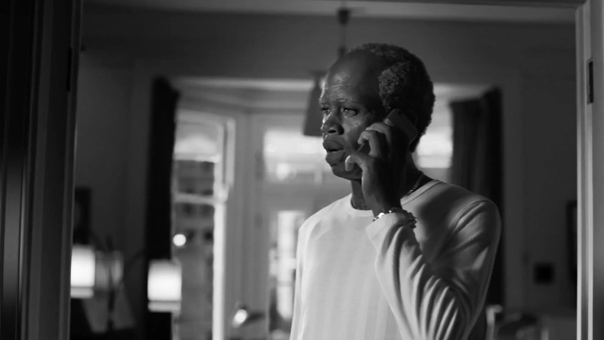An older Black man with short hair and a white long-sleeved shirt talks on a phone, looking concerned in a doorway of a home.