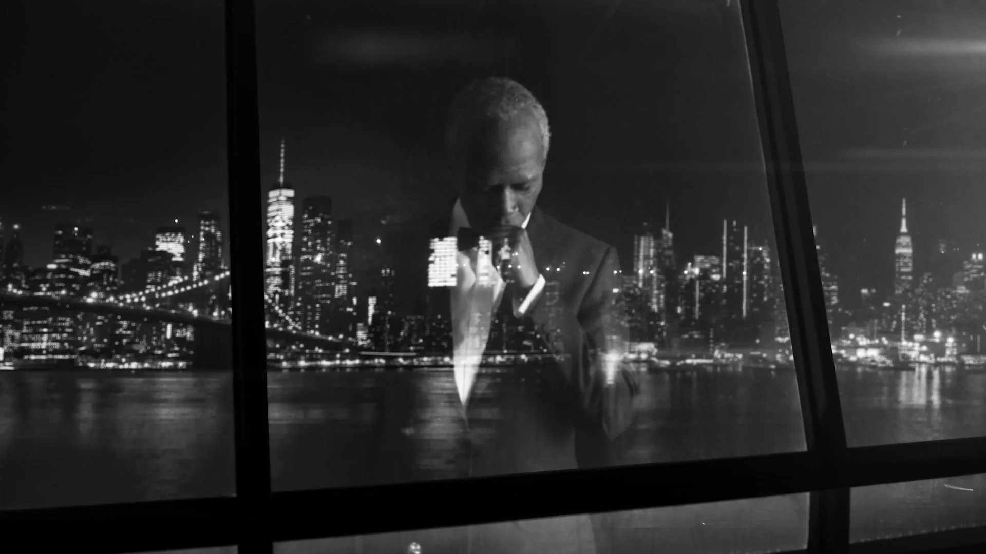 A distinguished older Black man in a tuxedo adjusts his bow tie, reflected in a window overlooking a sparkling city skyline at night.