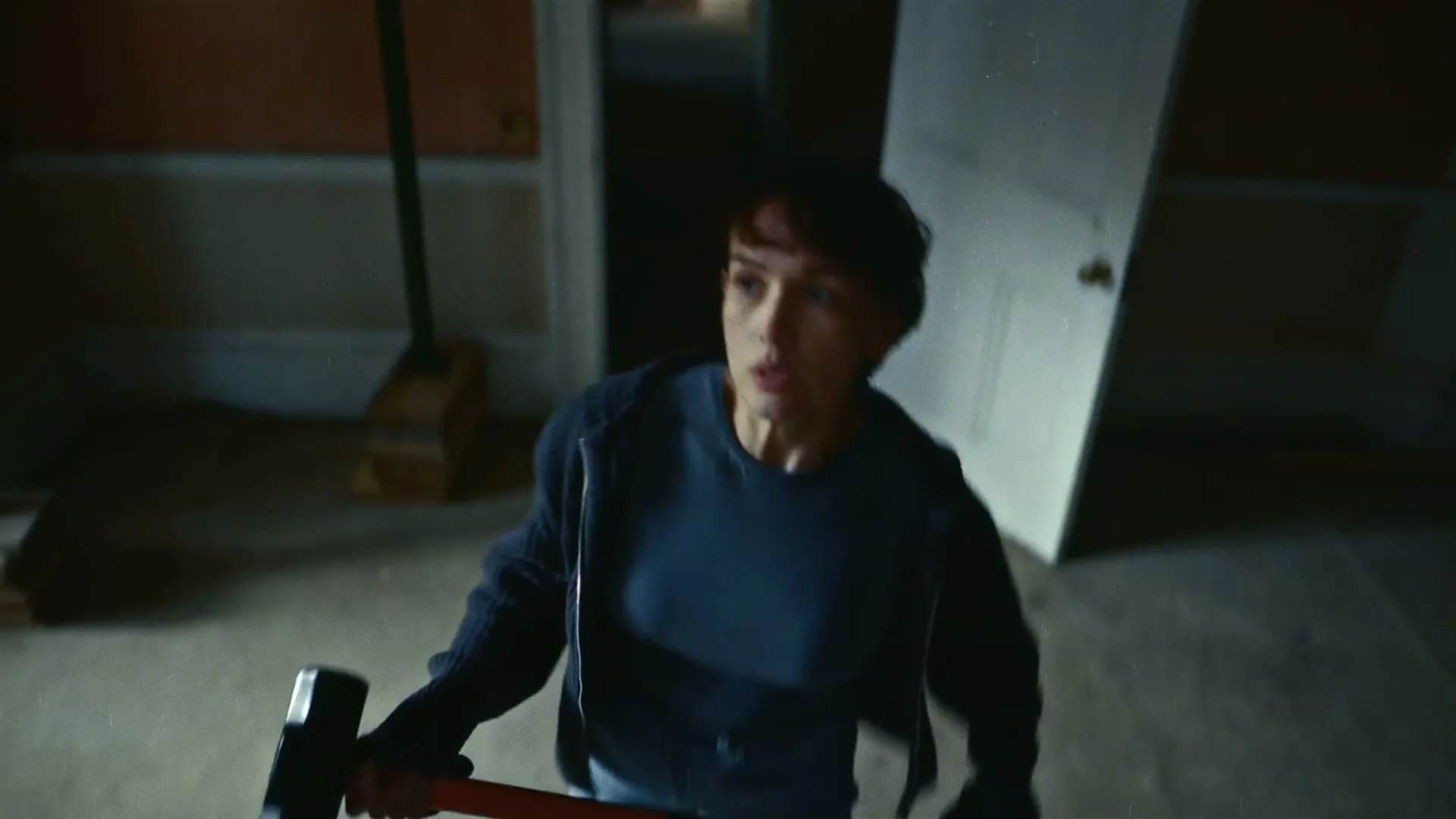 A woman with short brown hair, wearing a blue t-shirt and dark hoodie, holds a sledgehammer and looks around an empty, dimly lit room.