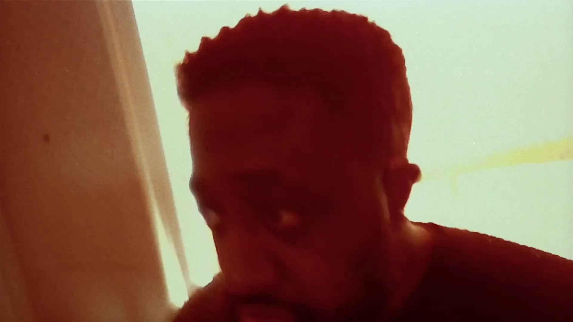 A man with dark skin and short hair looks up with a surprised expression, his face partially obscured by shadow in a dimly lit room.