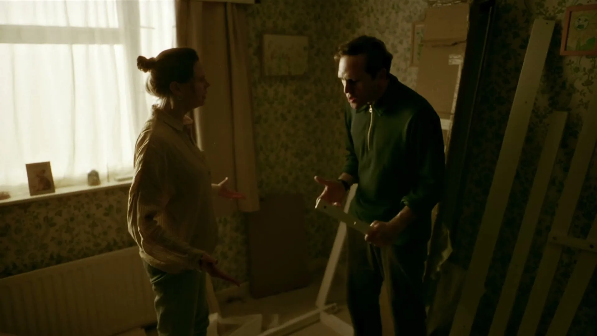 A man and a woman argue in a dimly lit room, surrounded by unassembled furniture parts, with sunlight streaming through a window.