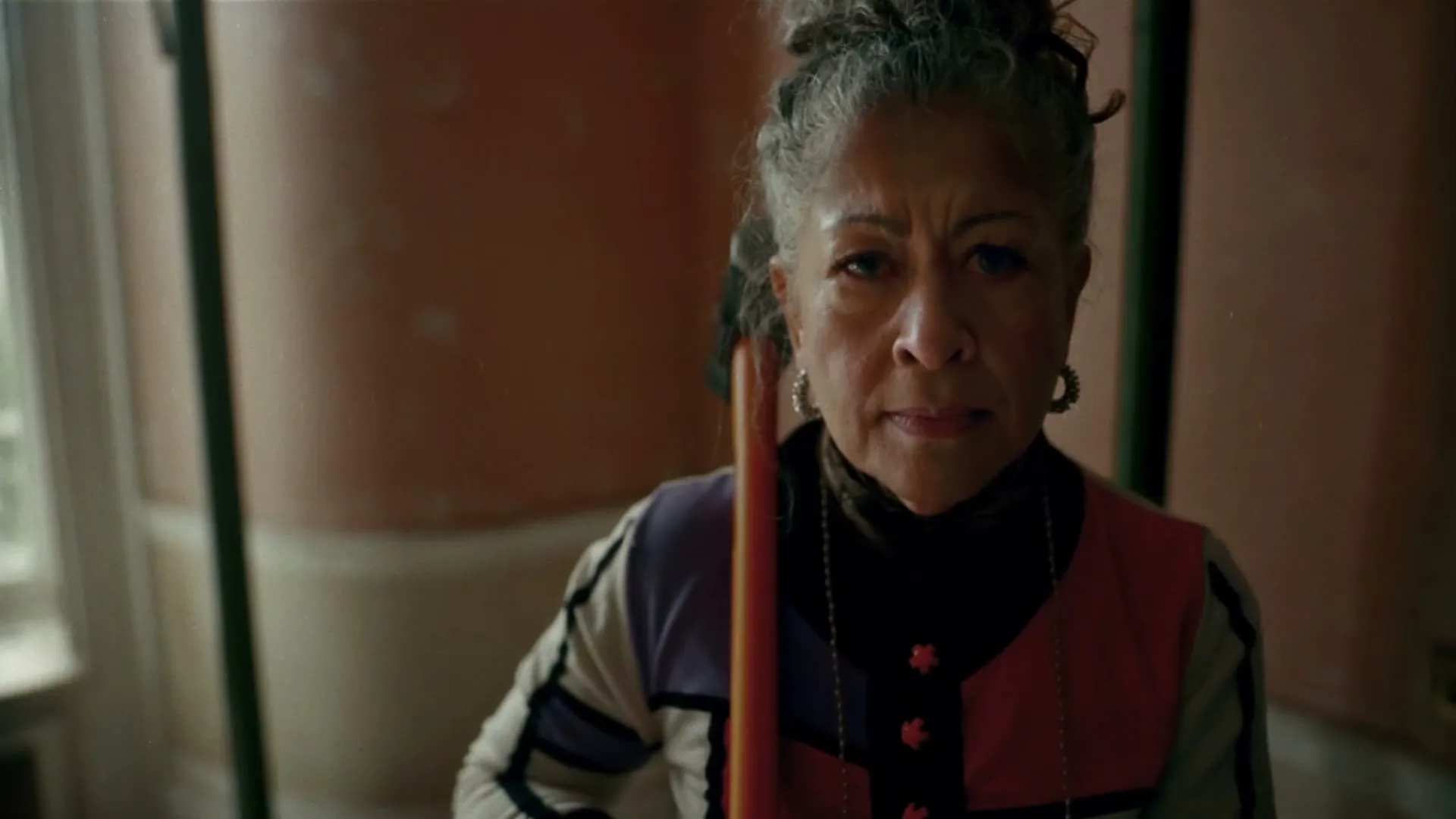 A serious older woman with grey hair and a colorful top holds a broom, looking directly at the camera in a room with a window.