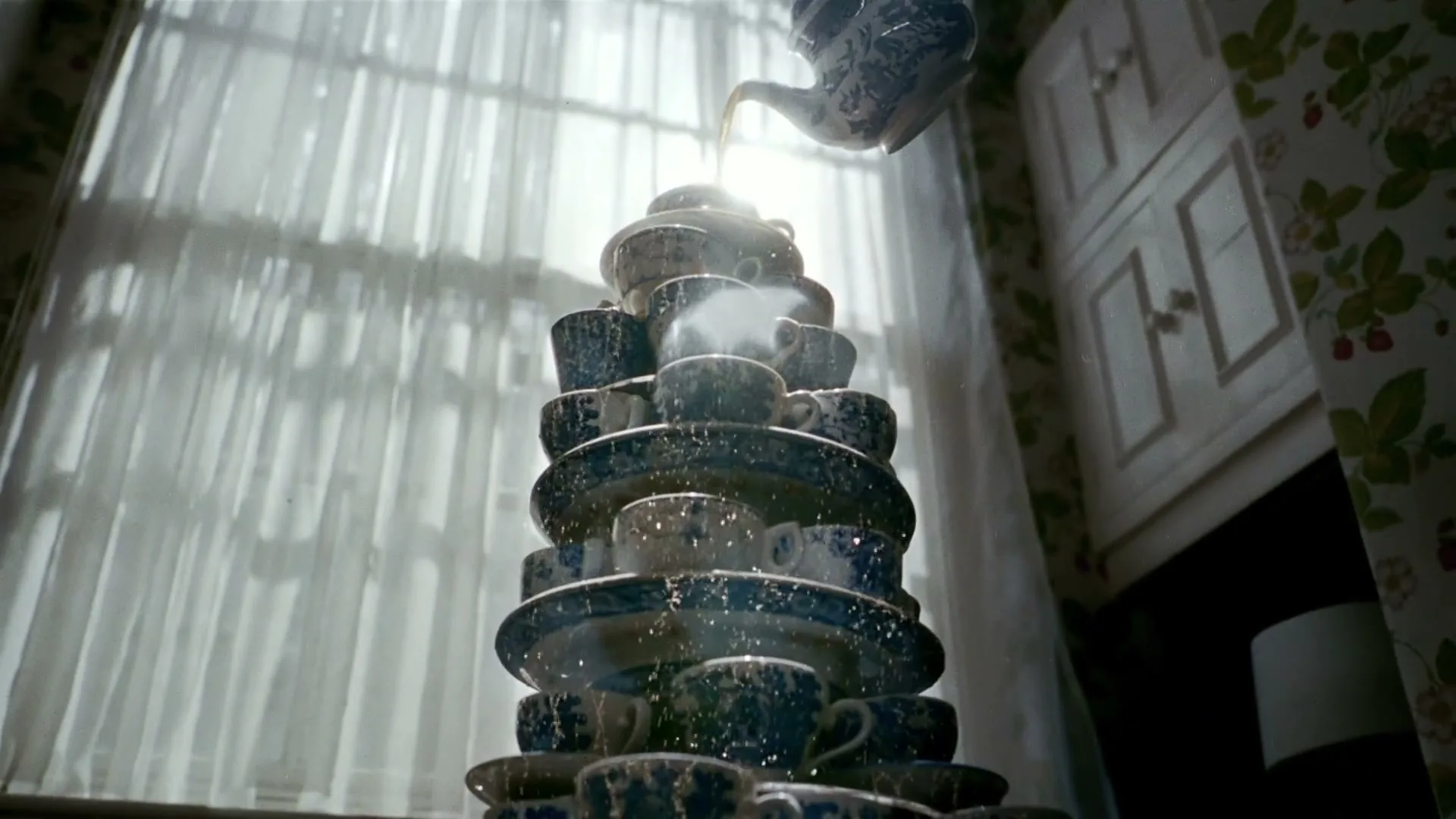 A blue and white teapot pours tea into a tall, precarious stack of matching teacups and saucers, creating a tea fountain in a sunlit room.