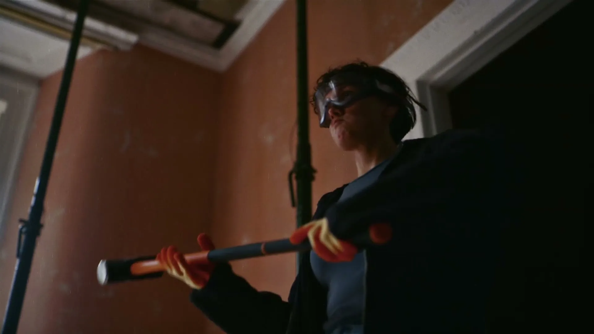 A person in safety goggles and work clothes swings a sledgehammer, breaking through a wall in a dimly lit room.