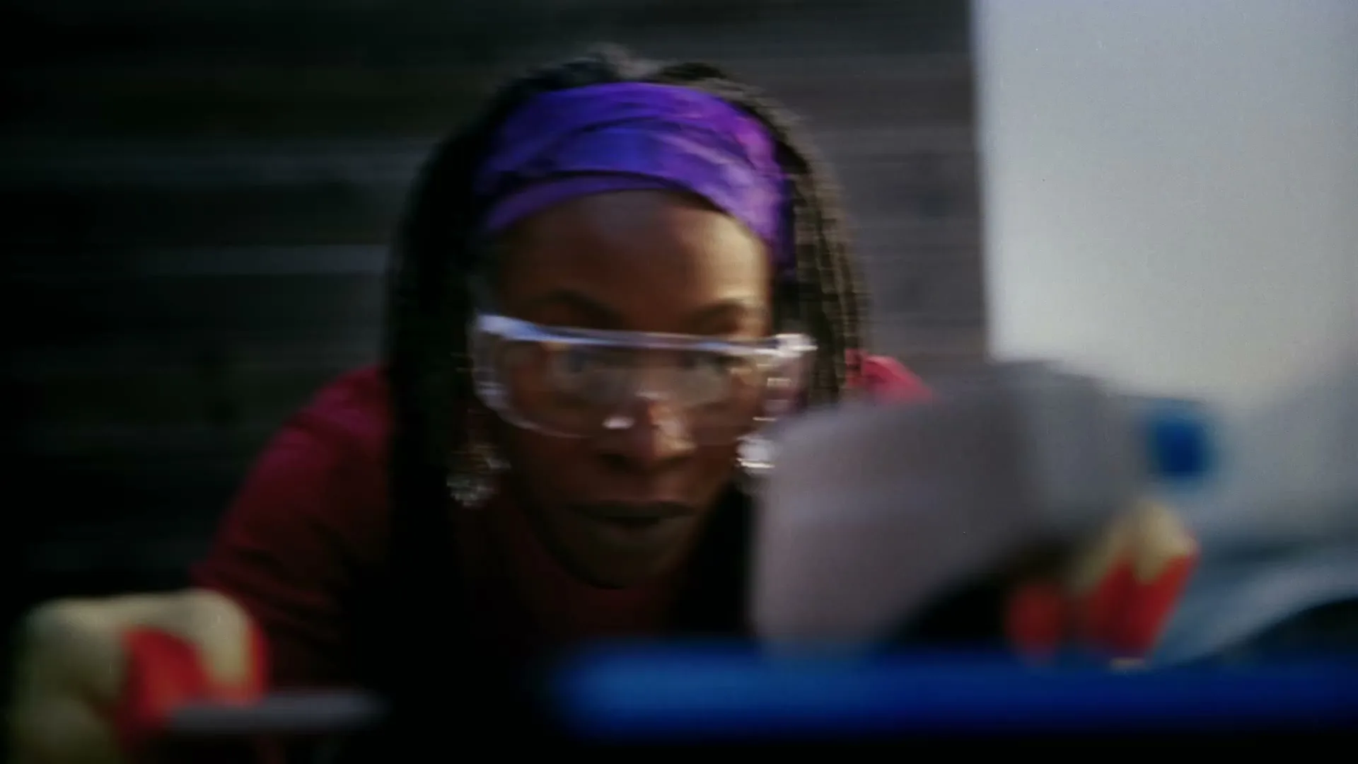 A woman with dreadlocks and a purple headscarf wears safety goggles and gloves while concentrating on a DIY project outdoors.