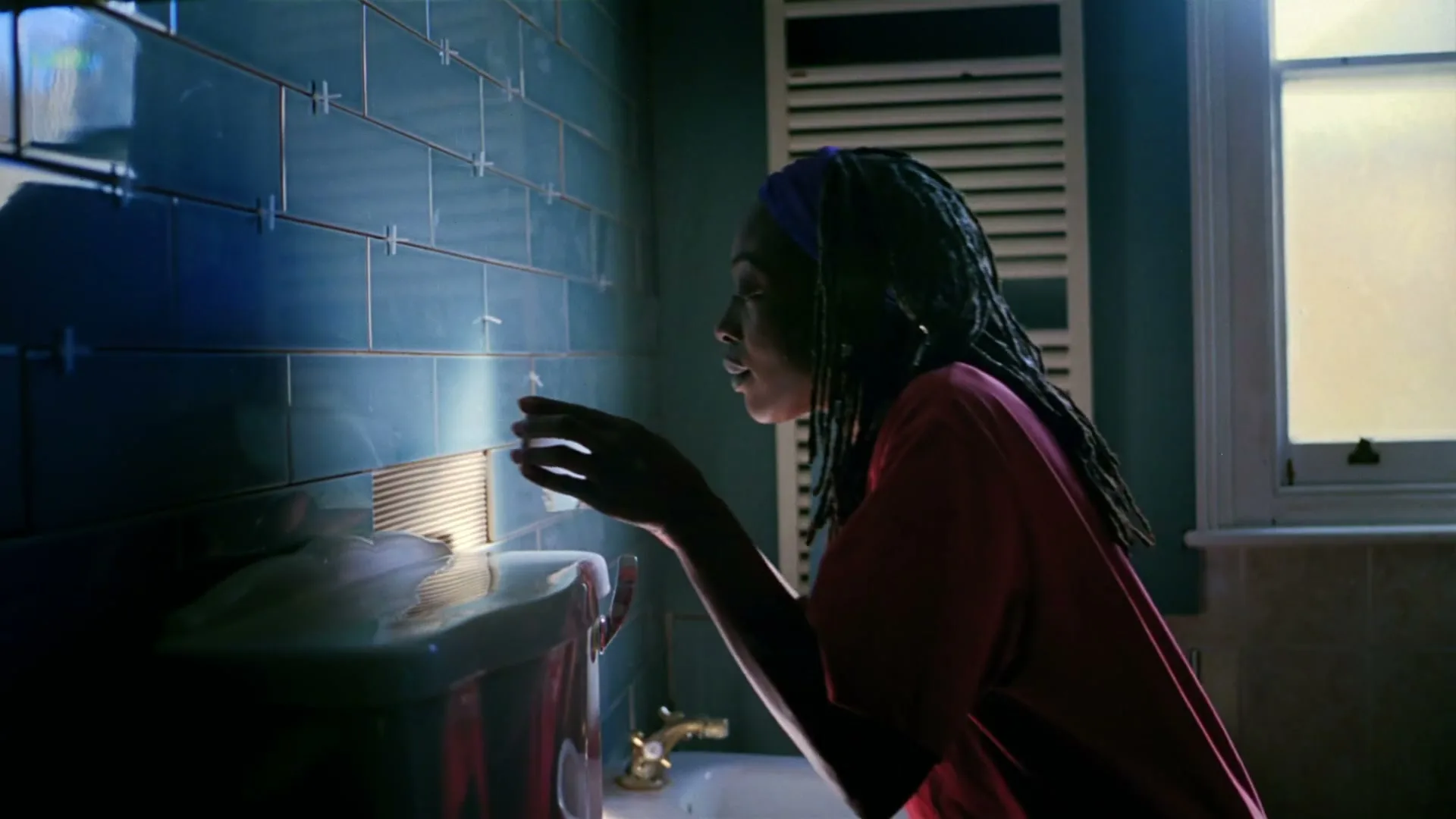 A young woman with dreadlocks and a purple headband looks at her reflection in a bathroom mirror, illuminated by a bright light.