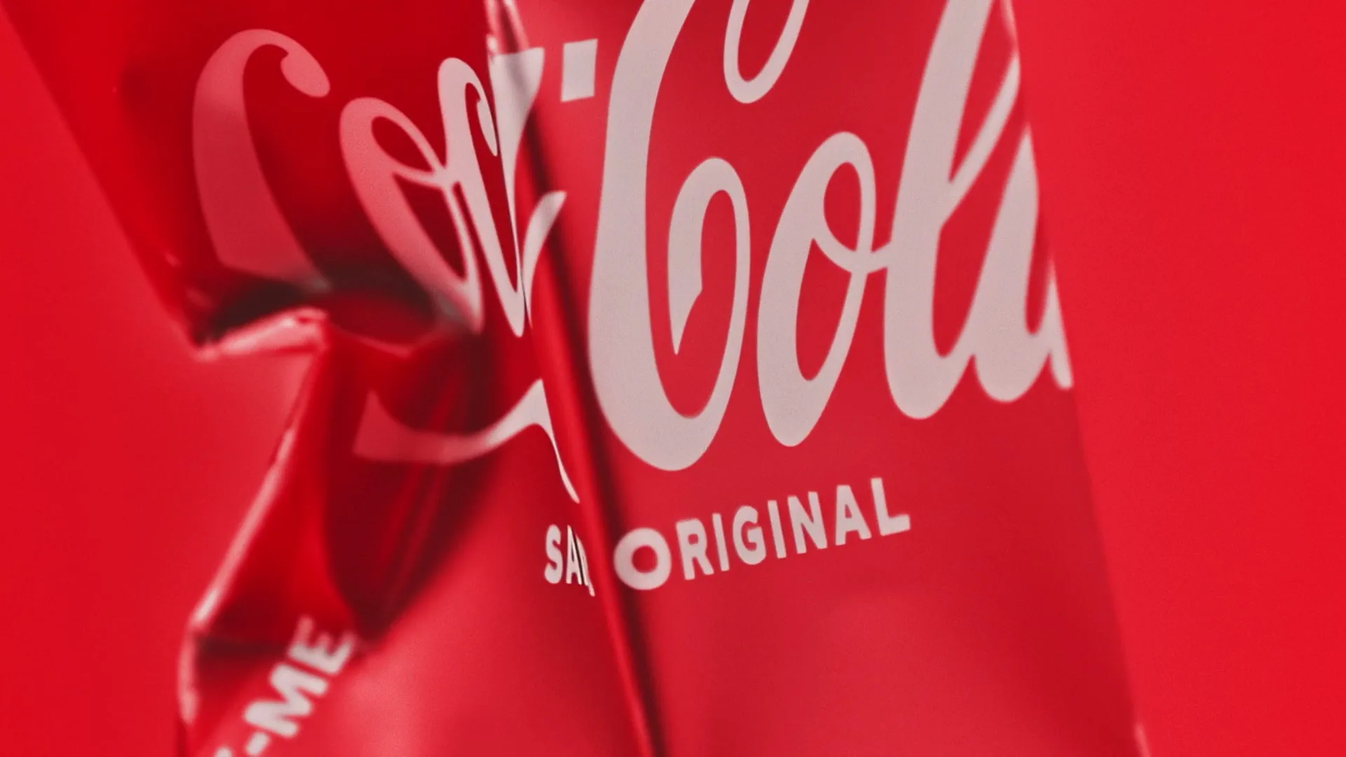 A close-up shot of two red Coca-Cola cans, one slightly crumpled, against a red background, with the logo and "SABOR ORIGINAL" visible.