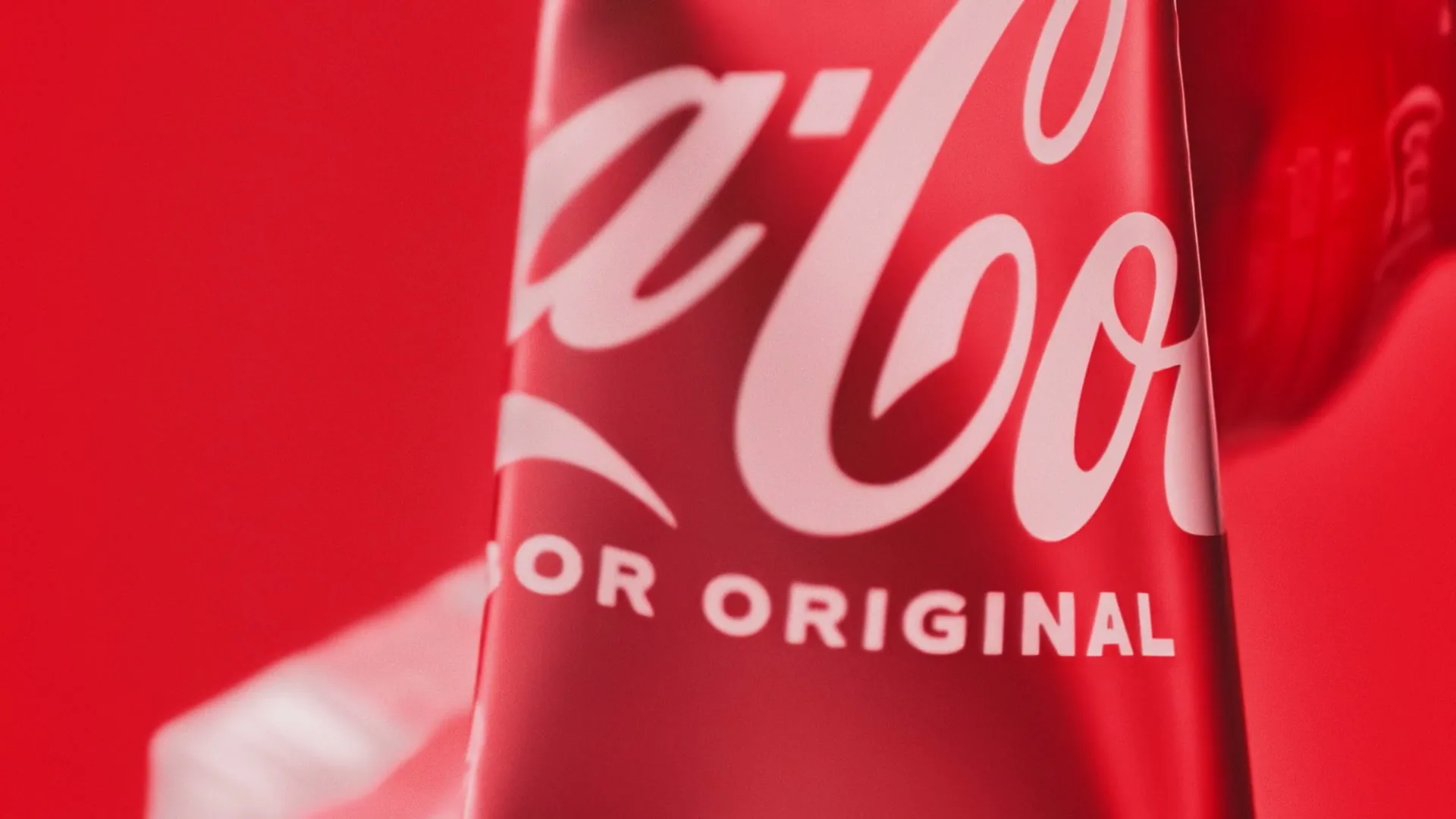 A close-up shot of a red Coca-Cola can with "OR ORIGINAL" visible, against a red background, with a hand in the background.