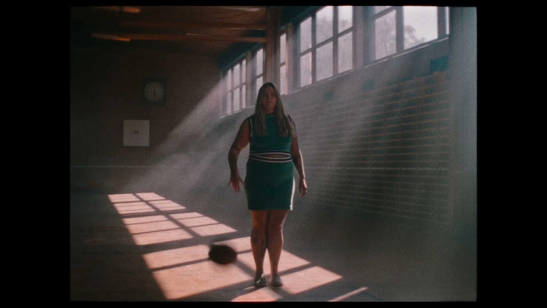 A woman in a teal dress stands in a sunlit gym, bouncing a basketball repeatedly, looking directly at the camera.