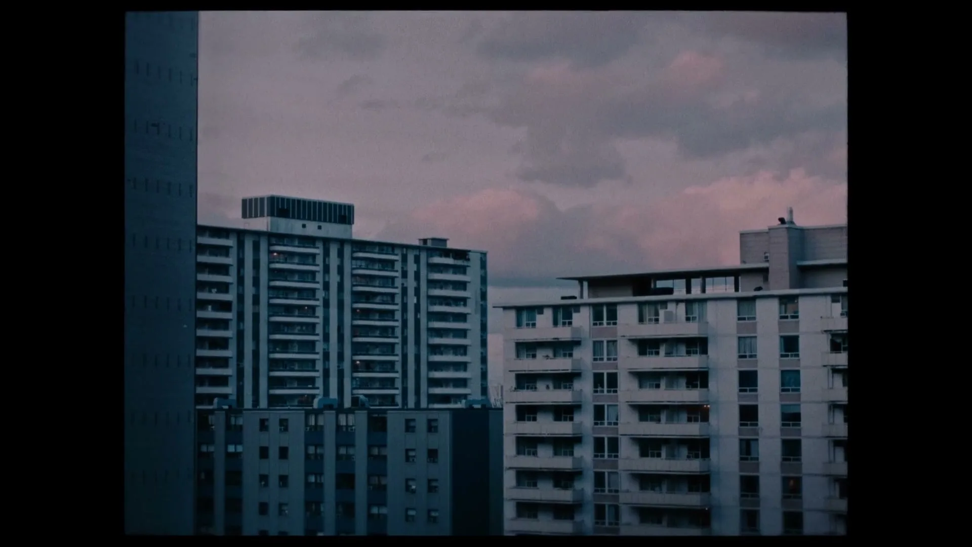A wide shot of several apartment buildings under a cloudy sky, with a subtle yellow film grain effect.