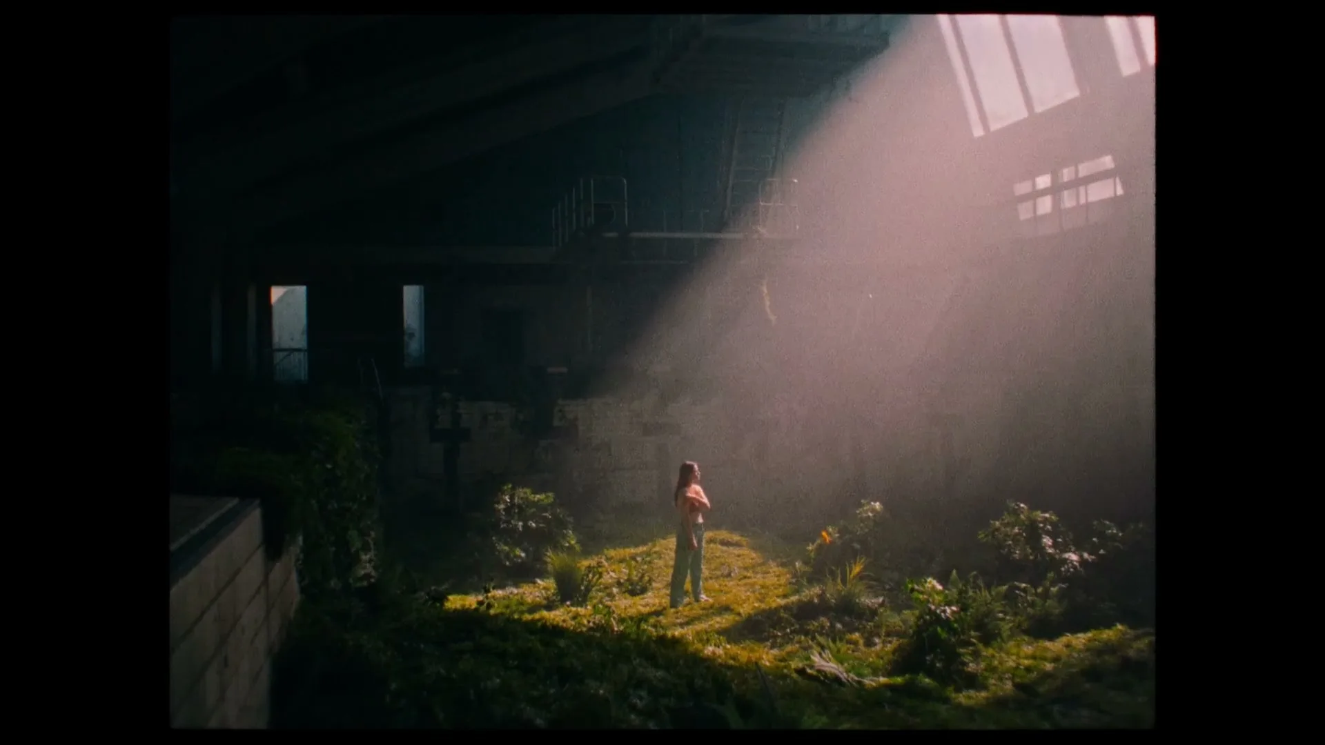 A young woman stands in a beam of light in a dark, overgrown abandoned building, looking around with a thoughtful expression.