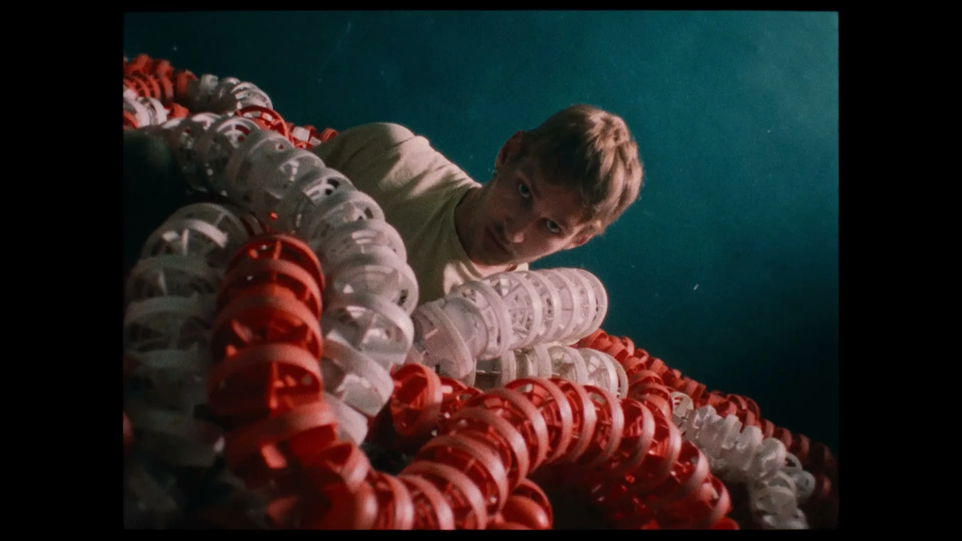 A young man with blonde hair looks down at the camera from above, surrounded by red and white swimming lane ropes in a pool.