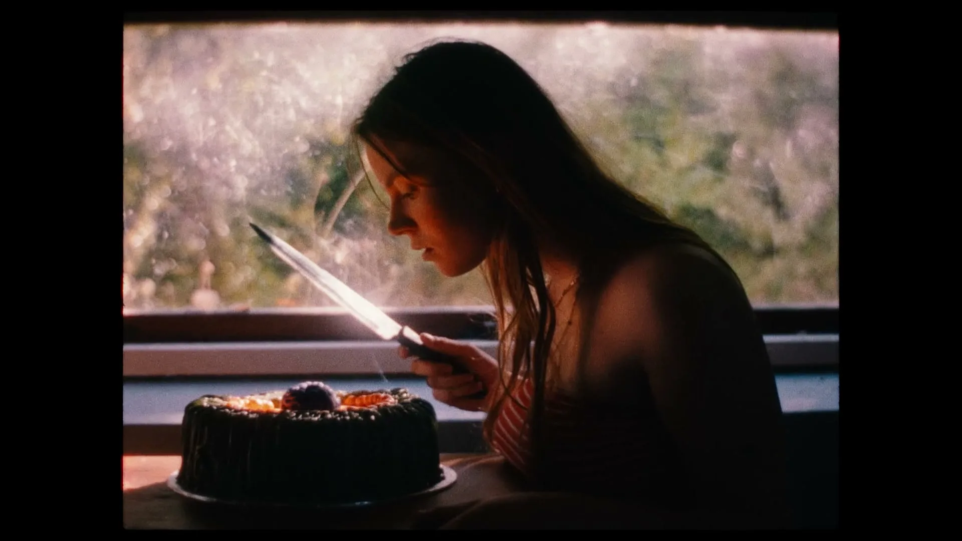 A young woman with long hair holds a knife over a cake, looking down intently, in a dimly lit room with a bright window in the background.