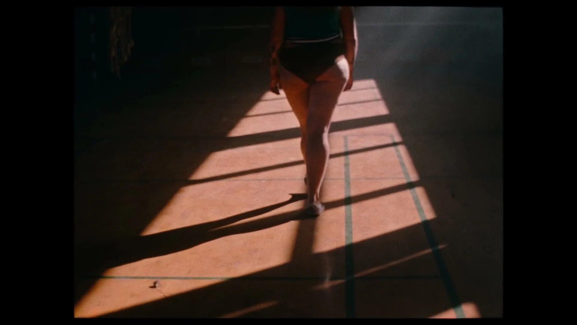A person in a swimsuit walks away from the camera across a sunlit gym floor, casting long shadows.