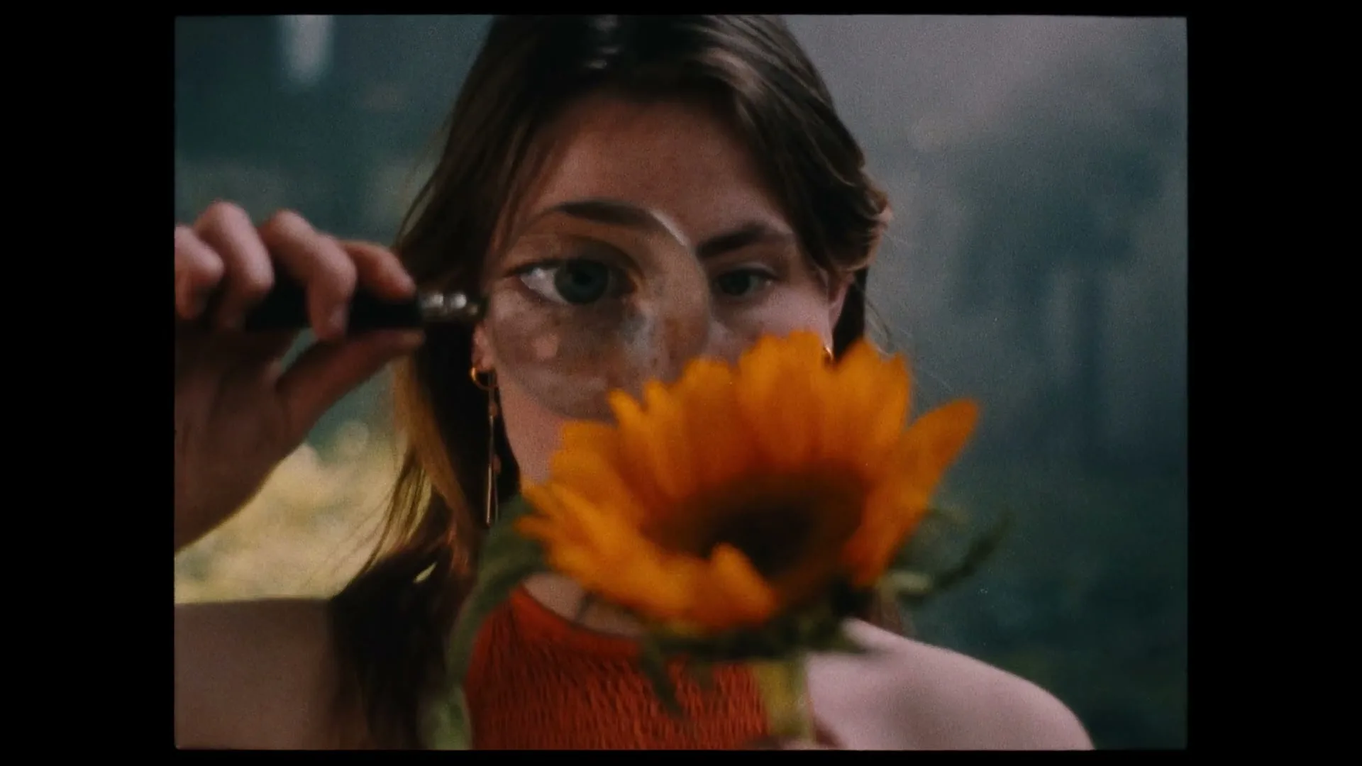 A young woman with long brown hair holds a magnifying glass to her eye, examining a sunflower in an outdoor setting.