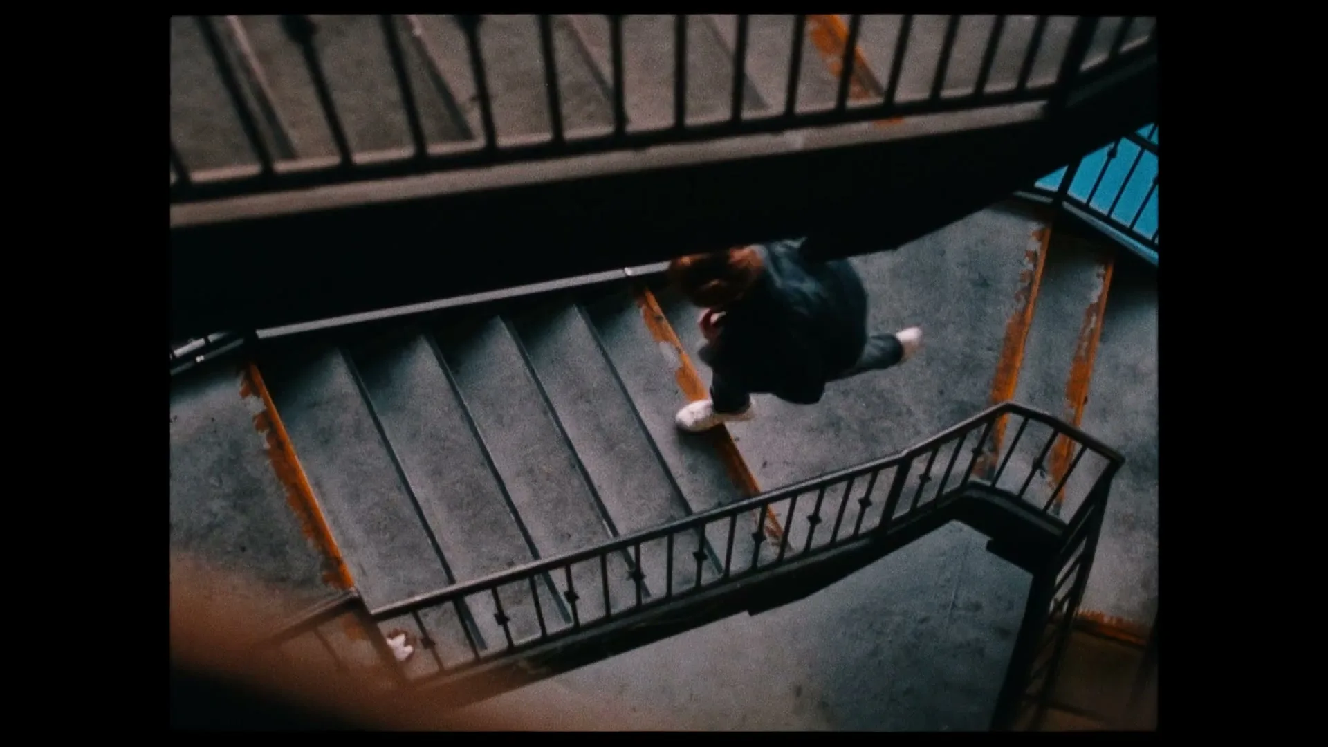 A person with red hair and a dark jacket walks down a concrete staircase with metal railings, viewed from an overhead perspective.