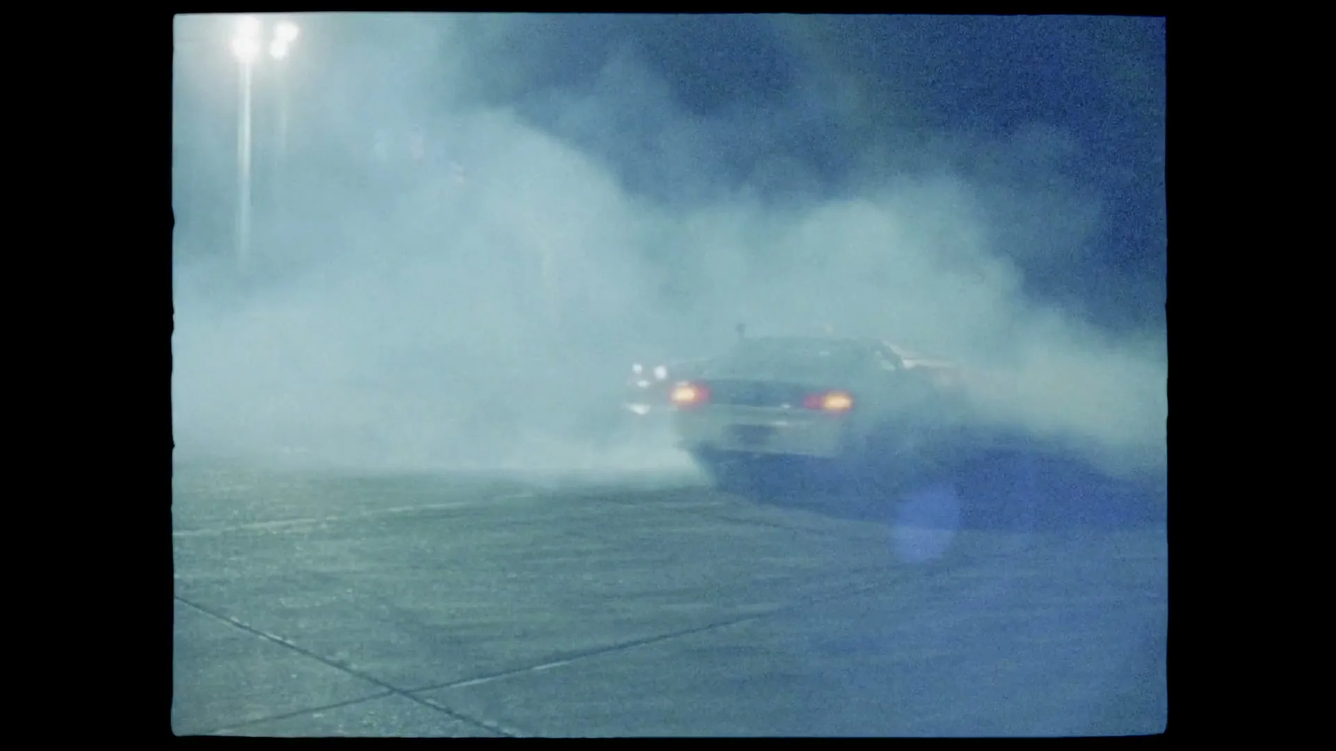 Two cars drift in a smoky parking lot at night, with bright lights in the background.