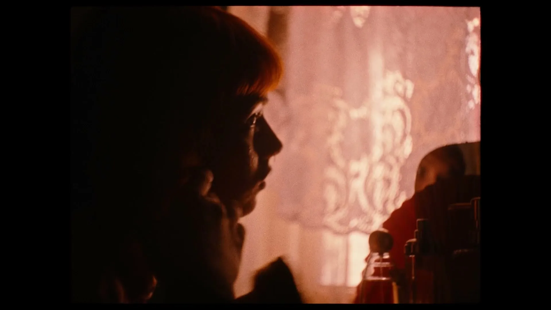 A young woman with red hair is seen in profile, looking out a window with a patterned curtain, in a dimly lit room.