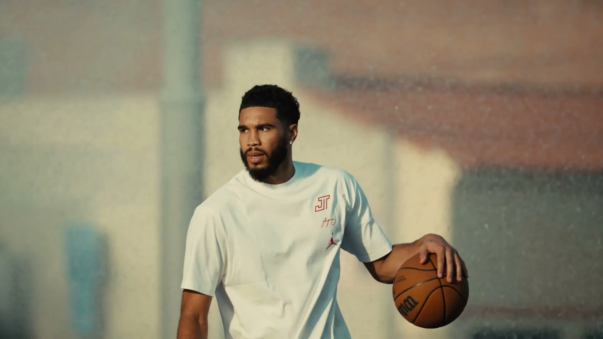 A male basketball player in a white t-shirt holds a basketball, looking intently to the side on an outdoor court.