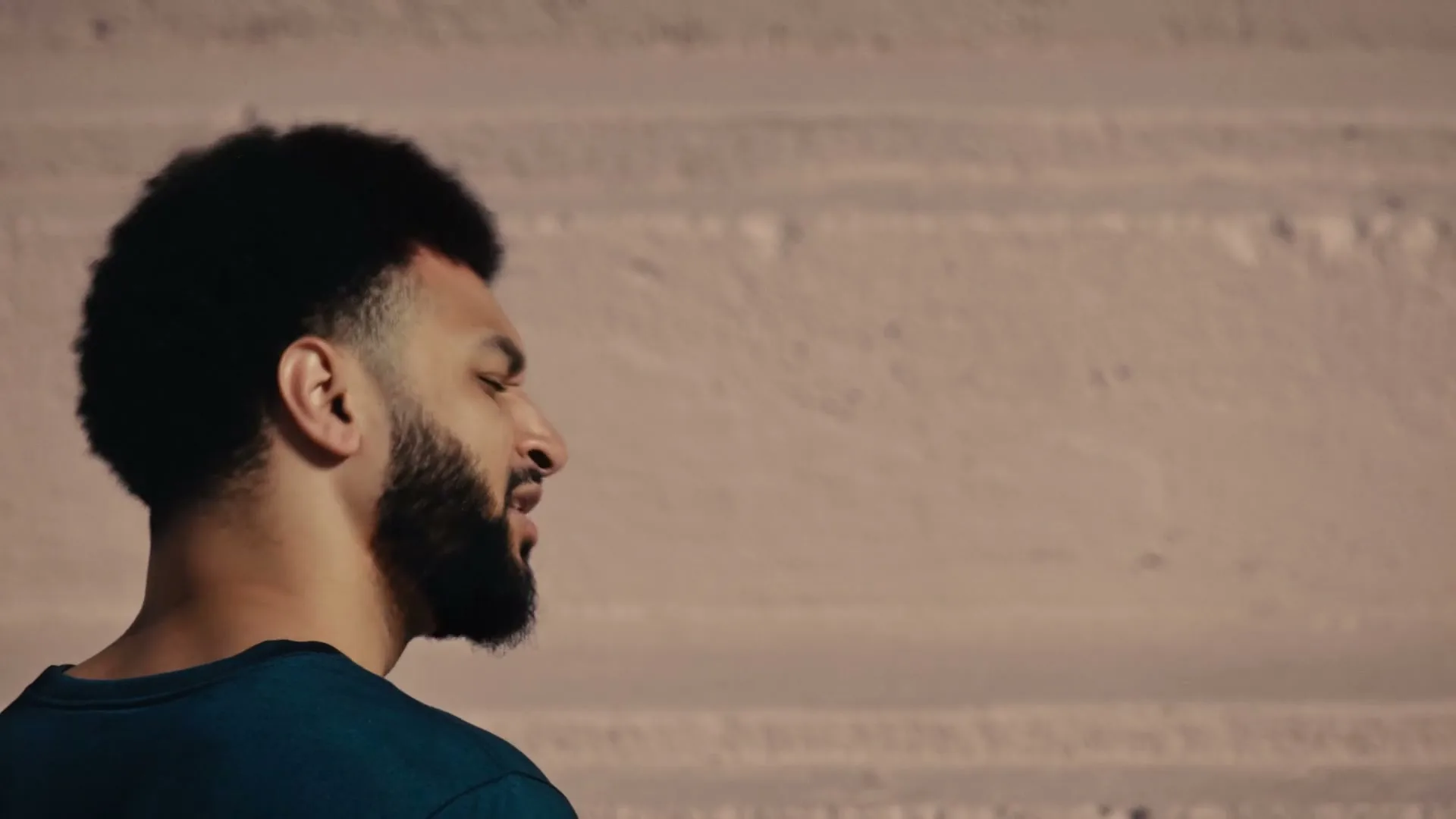 A man with a beard and dark curly hair looks right, with a light-colored wall in the background, bathed in warm sunlight.
