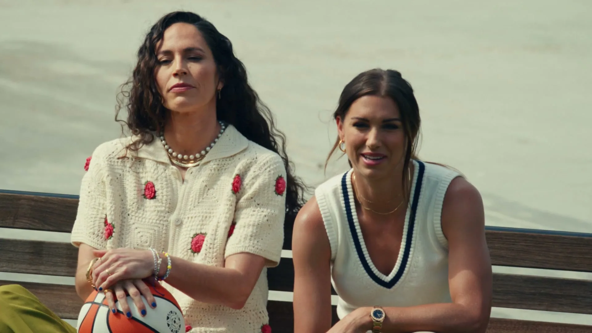 Two women, one with long dark hair and a cream crochet top, the other with a sleeveless cream top, sit on a bench outdoors.
