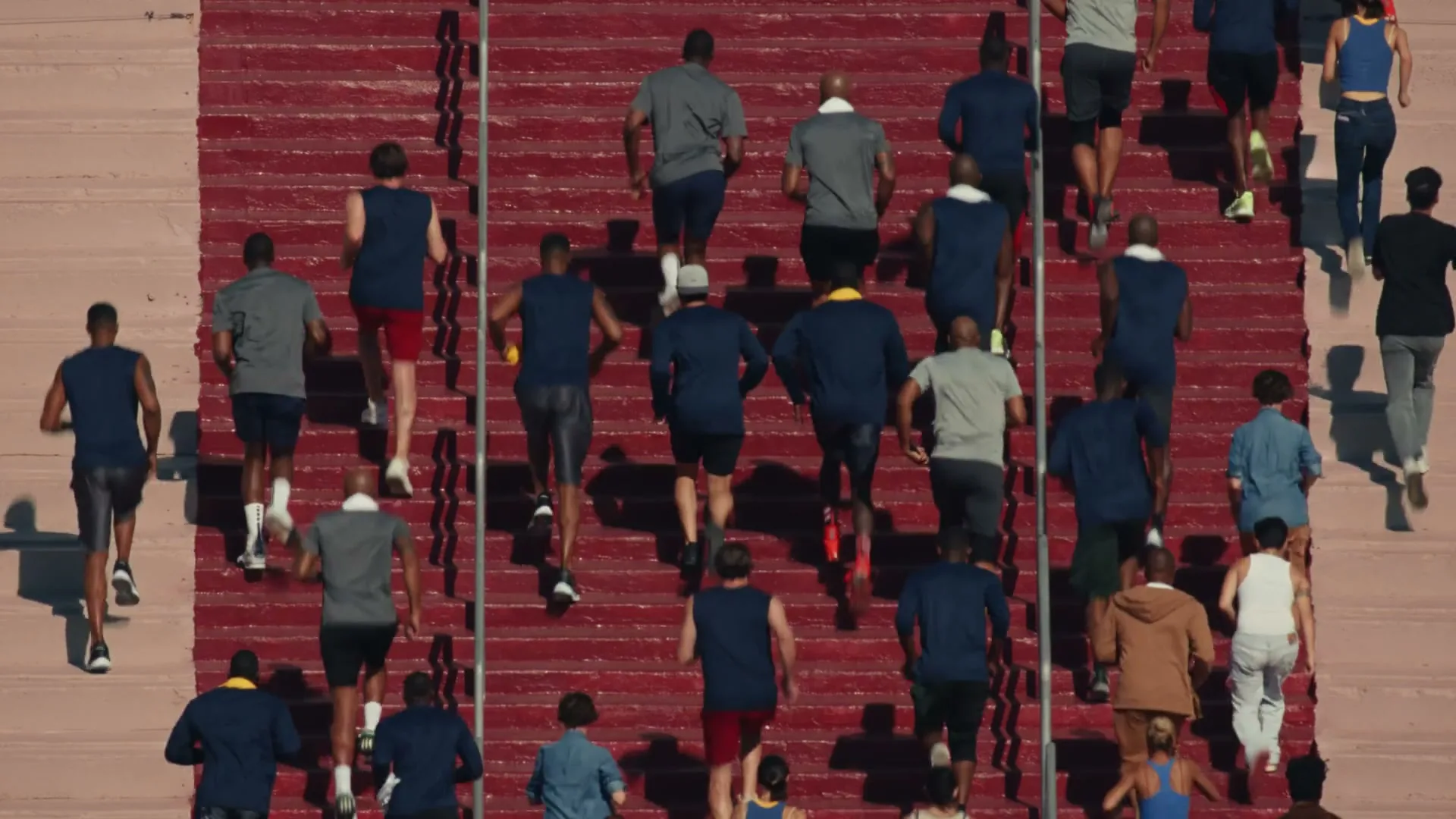 A diverse group of people of all ages and genders run up a wide, red stadium staircase, seen from an overhead perspective.