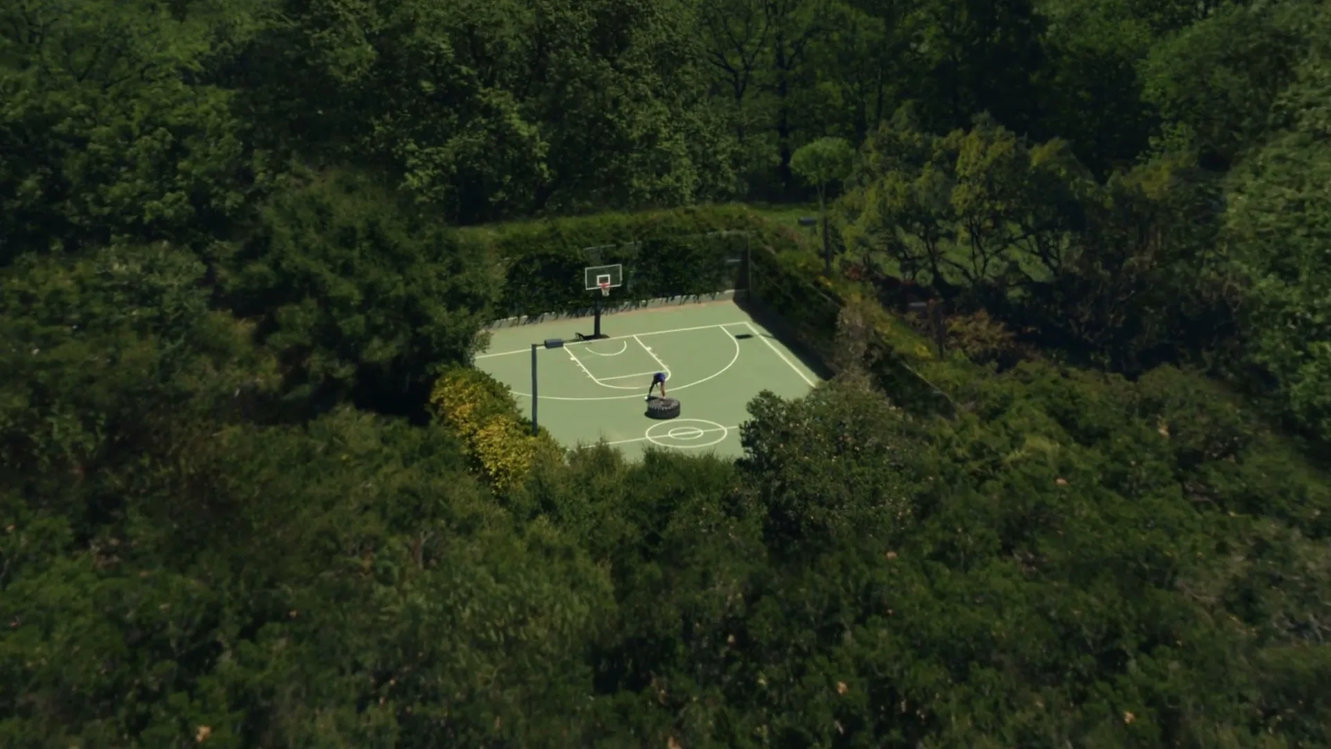 An aerial shot shows a male athlete in a blue shirt and black shorts flipping a large tire on a basketball court surrounded by lush green trees.