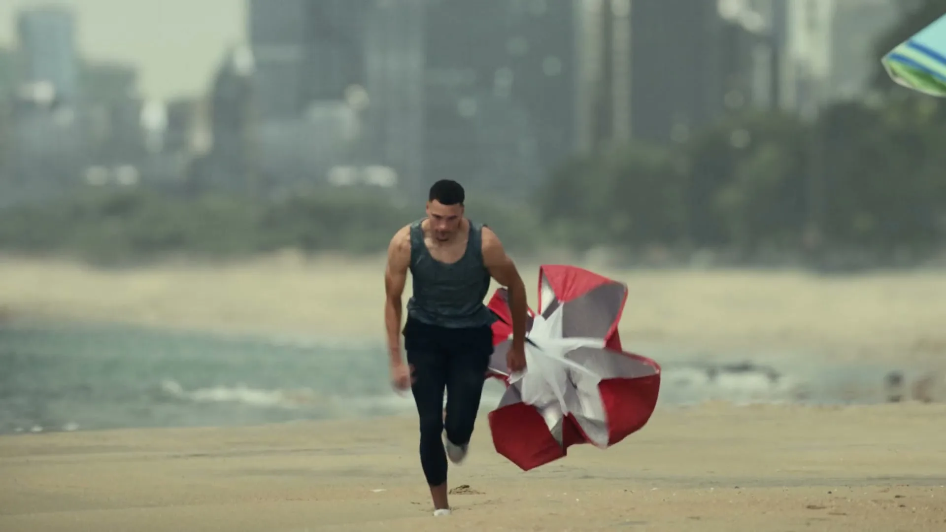 A male athlete runs on a sandy beach, pulling a resistance parachute behind him, with a city skyline in the background.