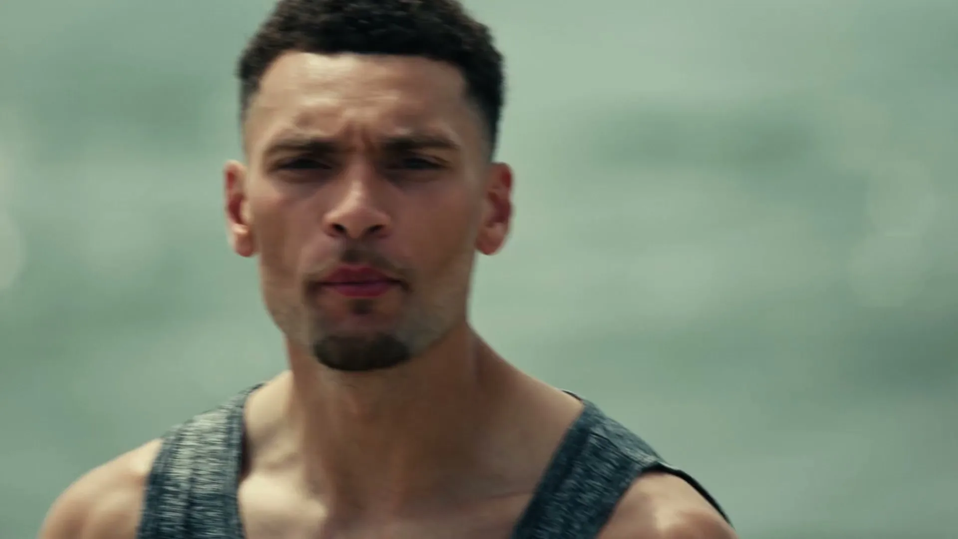 A male athlete in his 20s, wearing a grey tank top, shouts intensely at the camera with a blurred background.