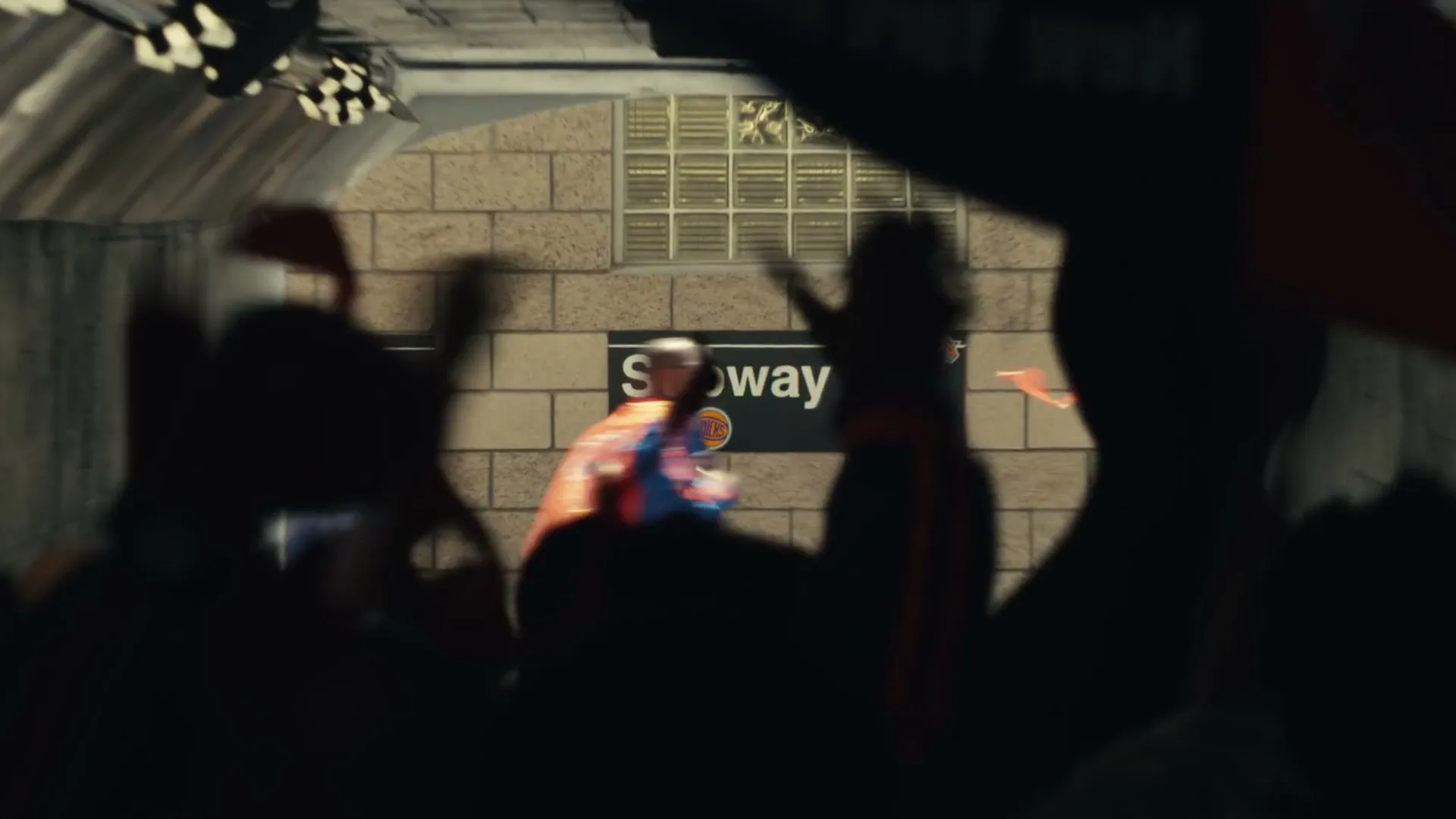 A crowd of silhouetted people with foam fingers and flags cheer as a man in a construction helmet and safety vest runs past a subway sign.