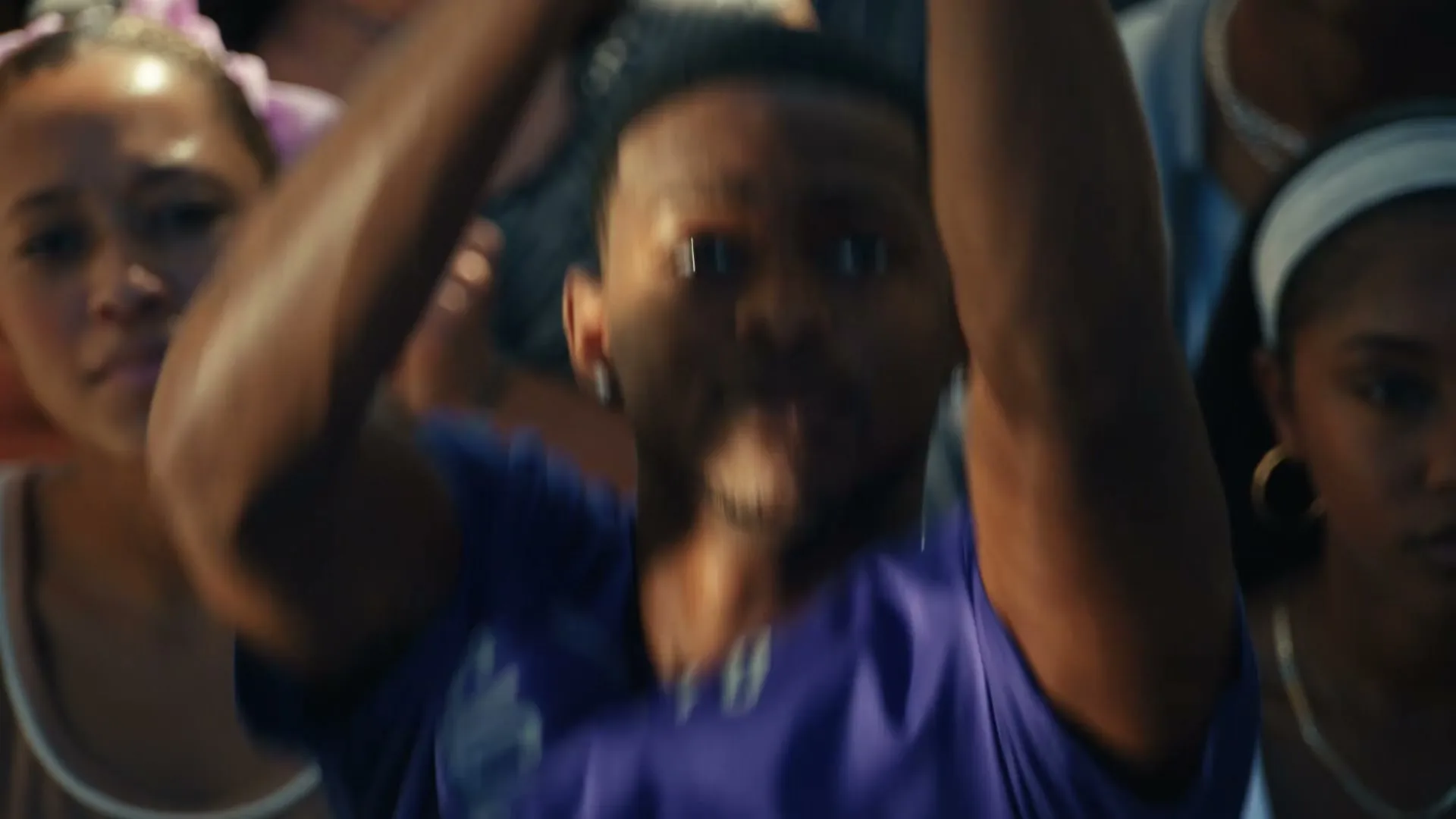 A young man in a purple shirt catches a basketball, then looks up with a hopeful expression, surrounded by other spectators at an outdoor game.
