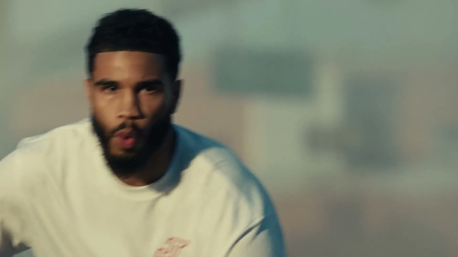 A young man with a beard and short hair, wearing a white t-shirt, looks intently at the camera while moving quickly.