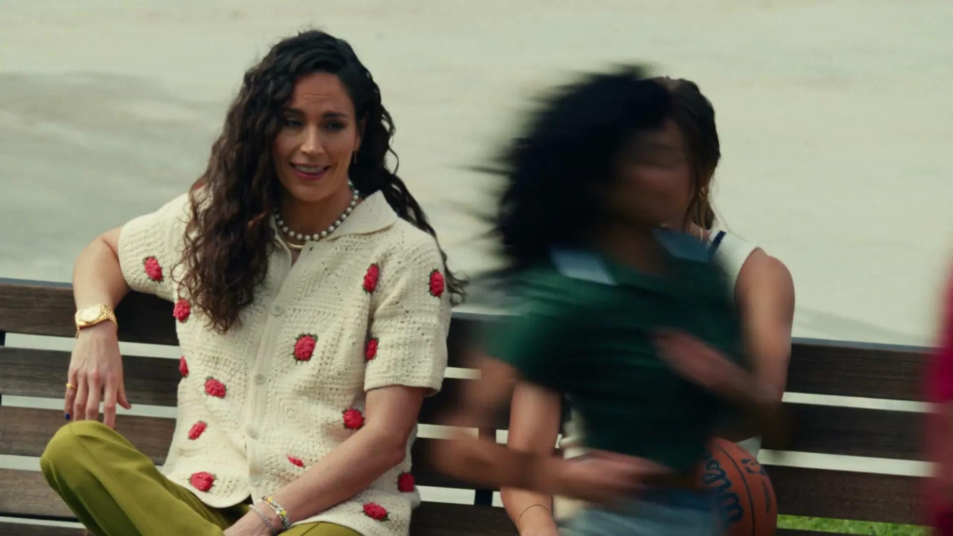 Two women, one with long curly hair and a strawberry cardigan, the other in a white v-neck, sit on a bench outdoors, smiling as people walk past.