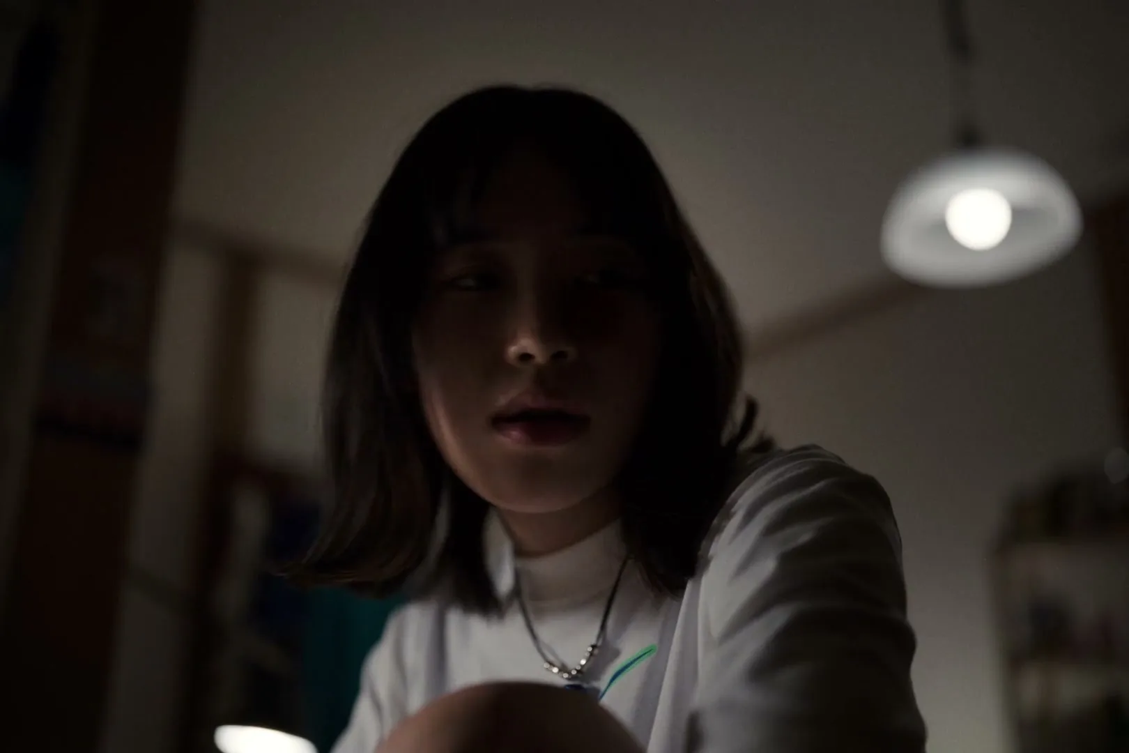 A young Japanese woman with short dark hair and a white t-shirt looks down with a concerned expression in a dimly lit room.