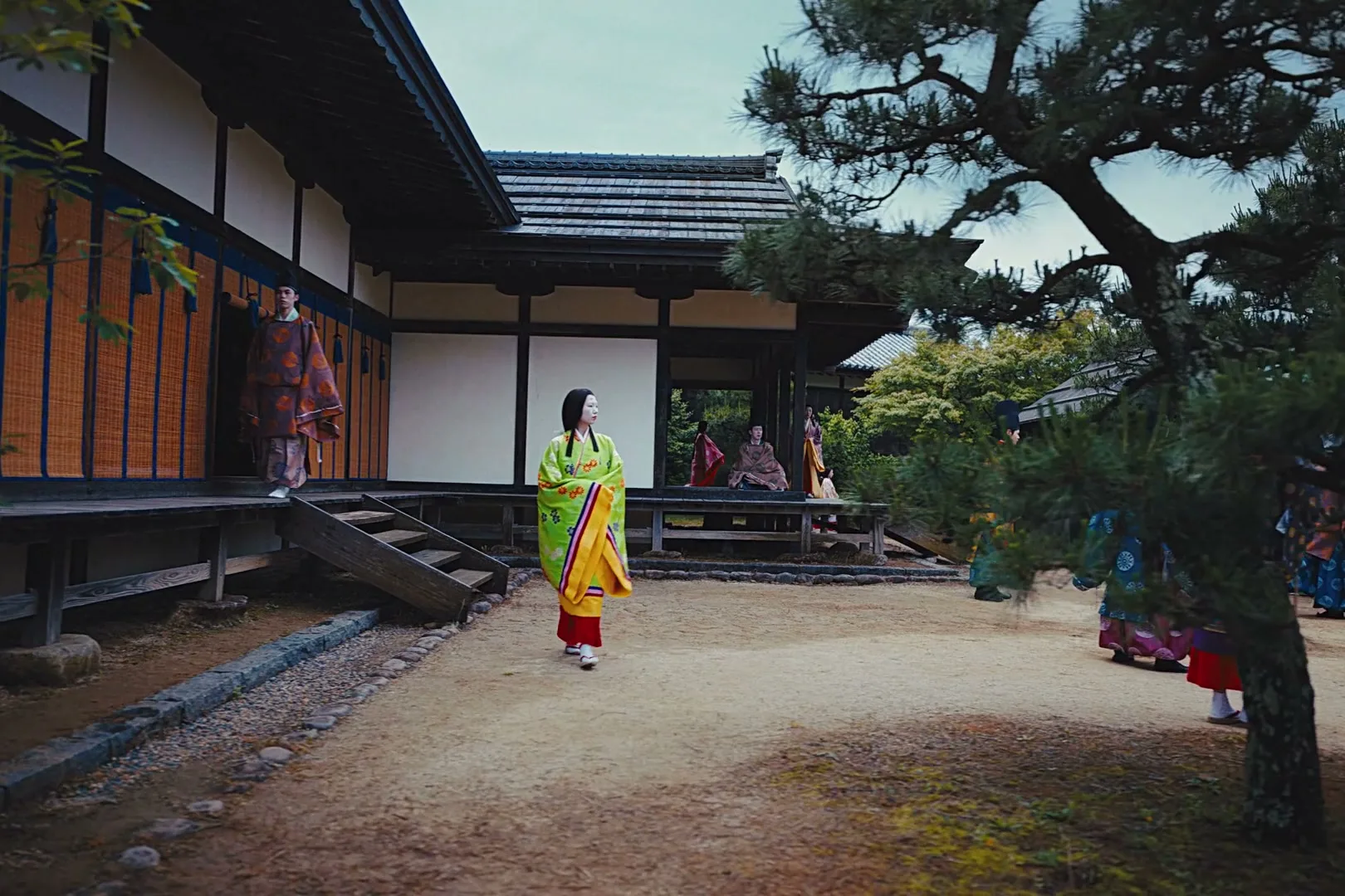 A woman in a bright green and yellow kimono walks across a dirt courtyard in front of a traditional Japanese building, while other people in kimonos are visible in the background.
